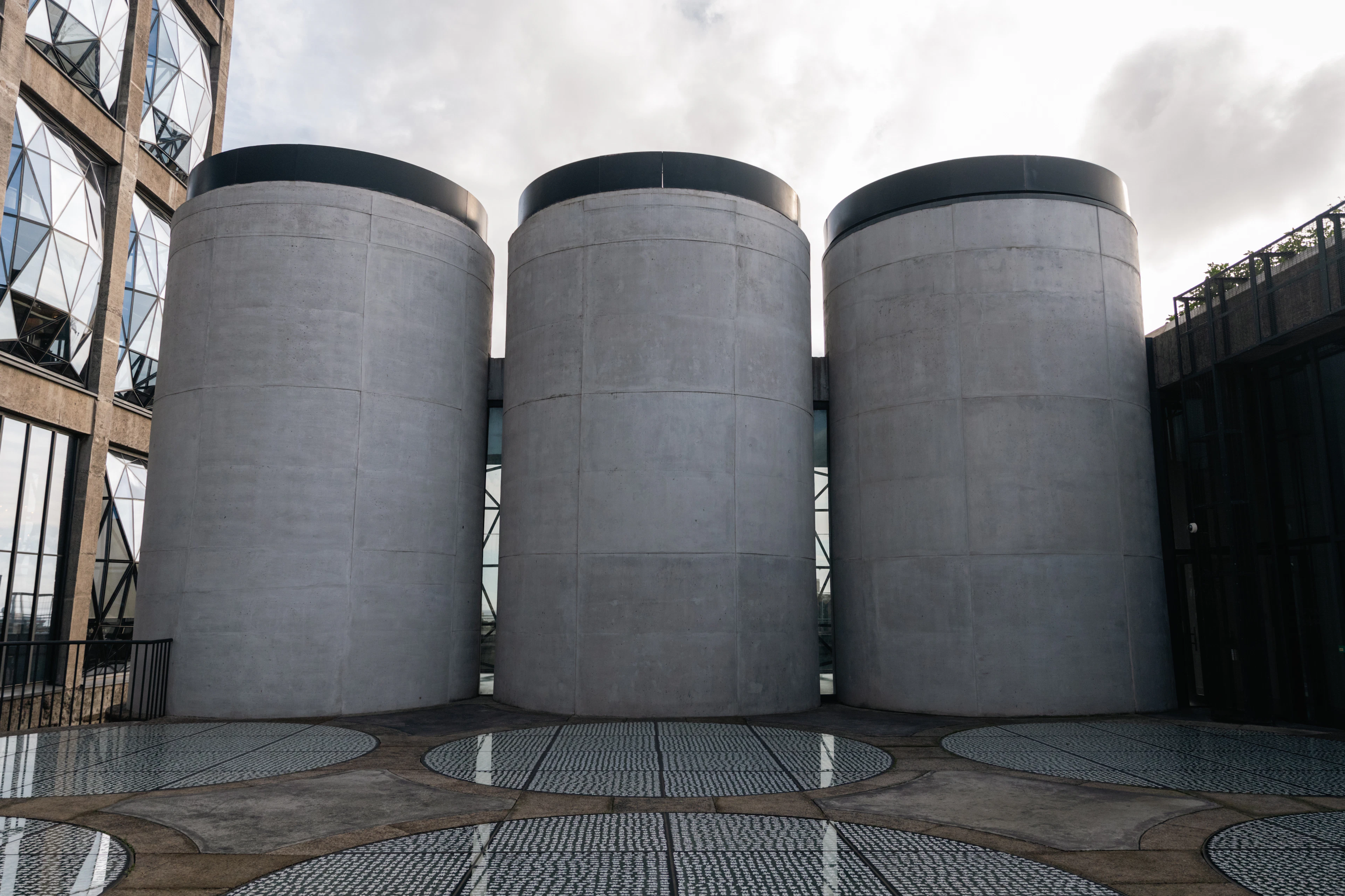 Three vast concrete grain silos standing on the rooftop terrace of the Silo Hotel at Cape Town's V&A Waterfront, beside the building's faceted pillowed-glass windows