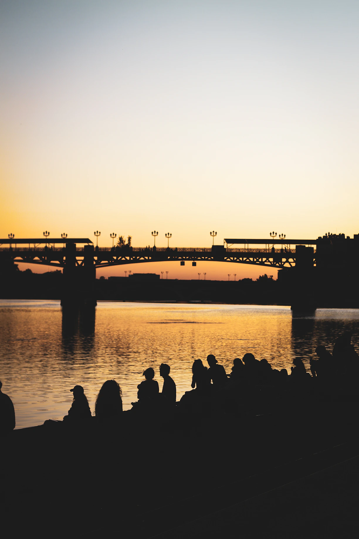 Silhouetted people watch sunset over river and bridge