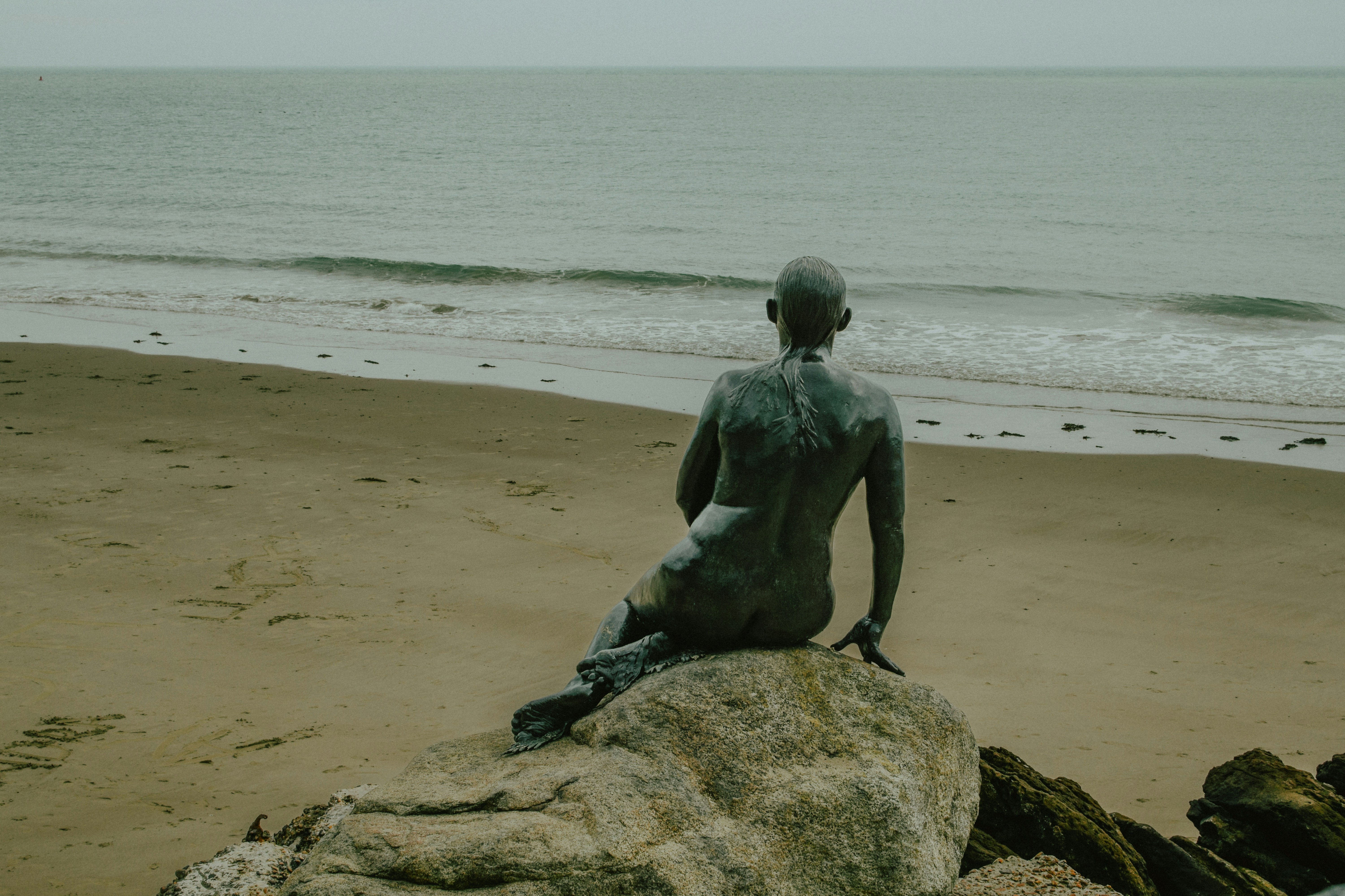 Statue einer Person, die auf einem Felsen am Meer sitzt.