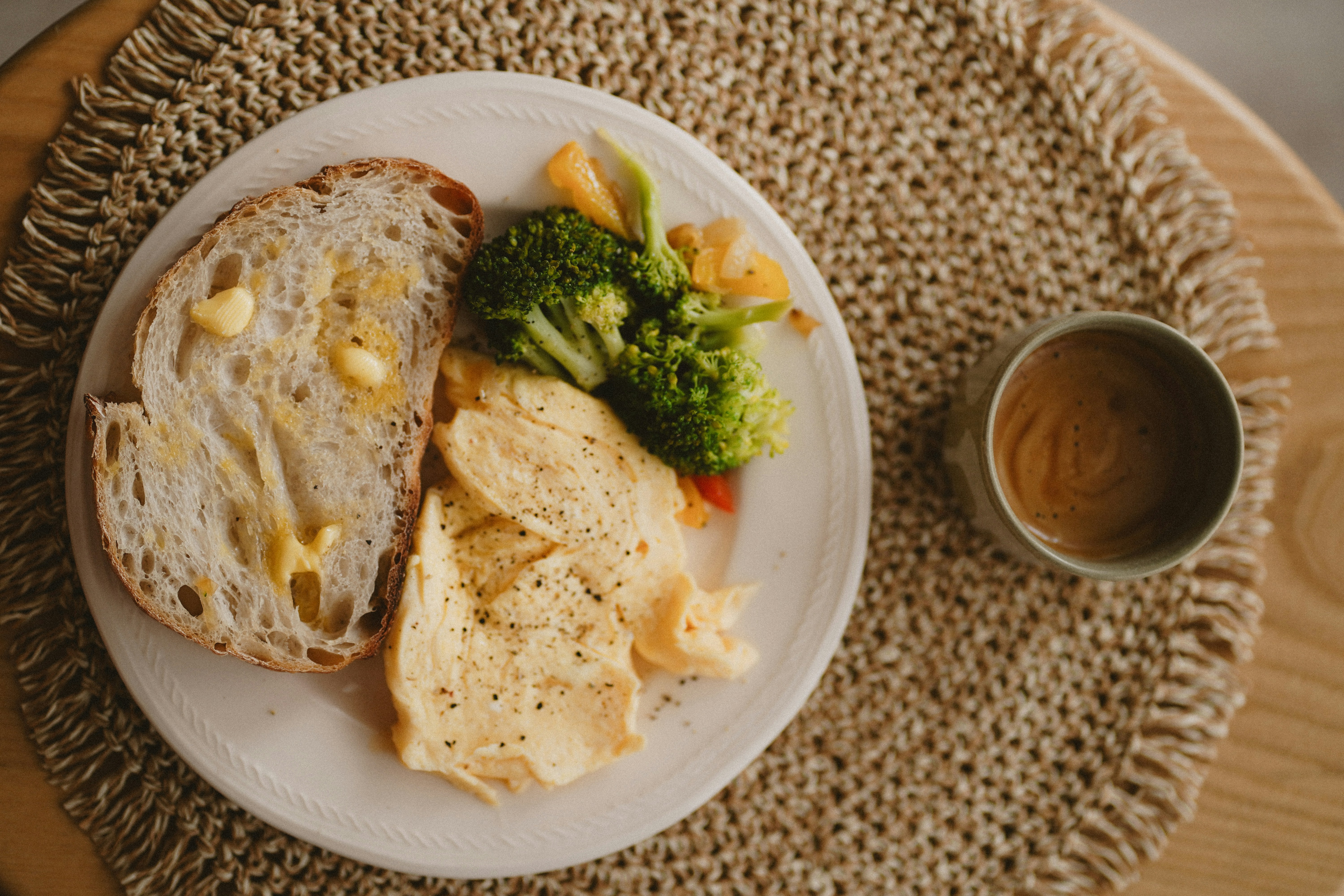 Warm, cozy breakfast setting with scrambled eggs, sautéed vegetables, buttered sourdough toast, and a ceramic mug of coffee on a woven placemat.