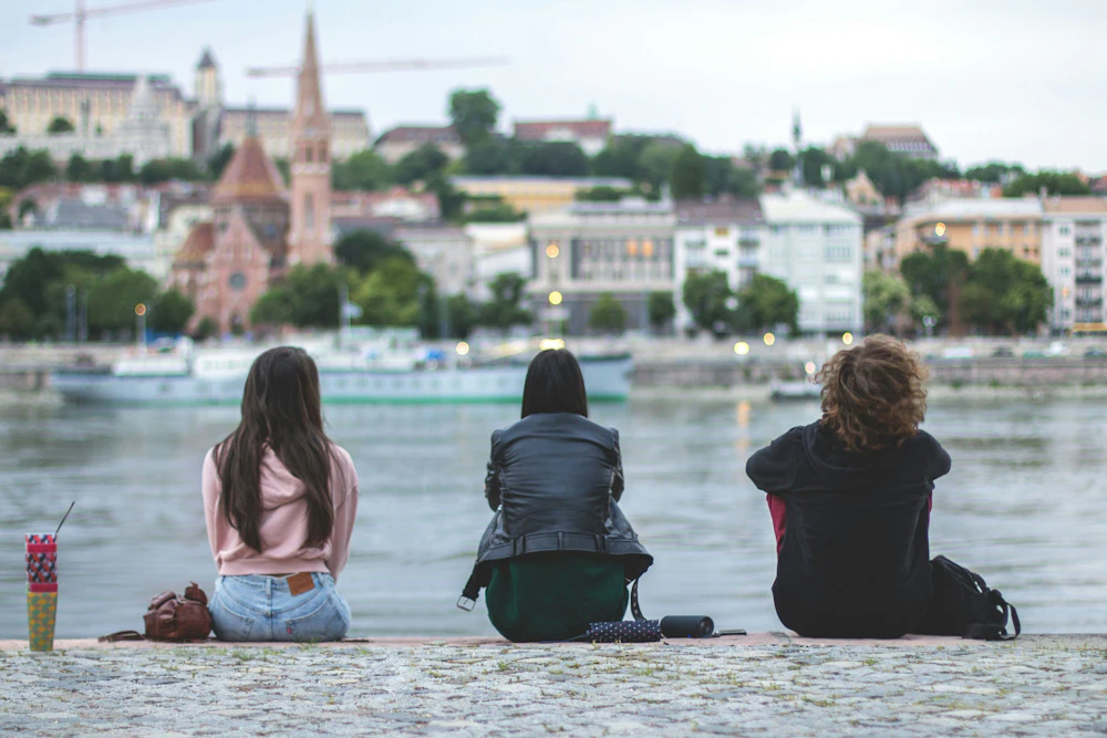 Three exchange students sitting by a river with a European city behind them