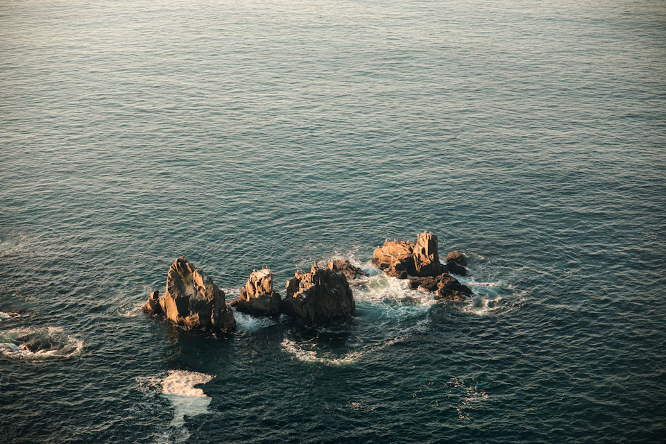 The image depicts a serene ocean scene with three large rocks jutting out from the water, creating a natural island amidst the vast expanse of blue sea. The rocks are positioned at different angles and heights, adding depth to the composition. The sky above is a light beige color, providing a beautiful contrast to the dark blue of the ocean below.