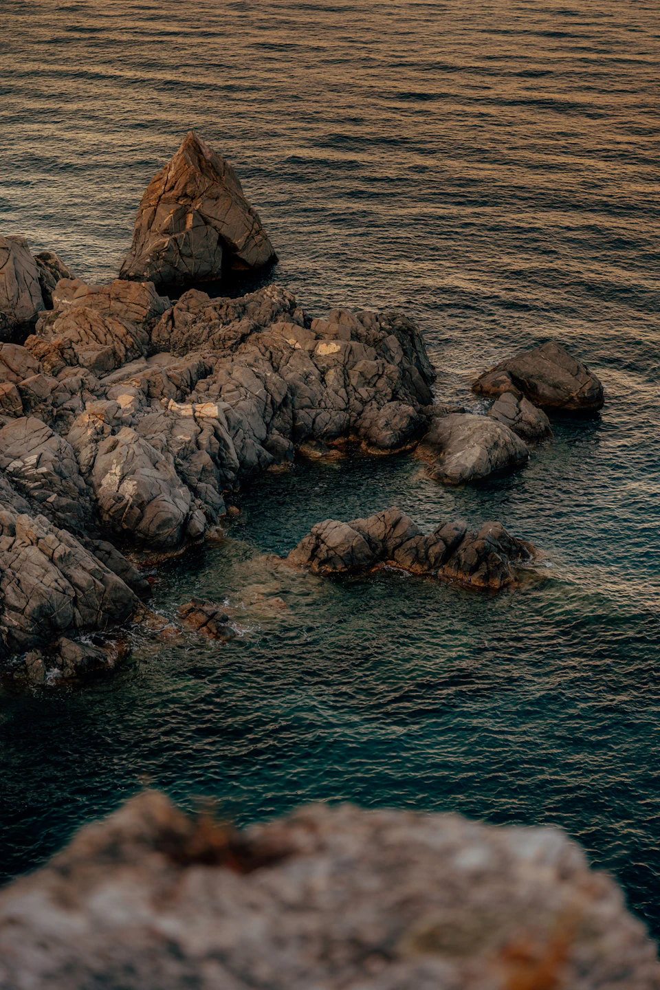The image depicts a rocky coastline with a large, jagged rock formation on the left side and smaller rocks scattered across the right side. The water surrounding the rocks is a deep blue color, suggesting it could be a body of water such as an ocean or sea. The sky in the background appears hazy, adding to the overall atmosphere of the scene.