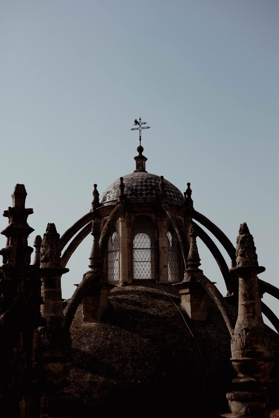 The image depicts a stone tower with a dome and cross on top, situated against a clear blue sky. The tower is adorned with numerous statues and carvings, adding to its majestic appearance. A weather vane is mounted atop the tower, indicating the direction of the wind. The perspective of the photo is from below, looking up at the tower, emphasizing its height and grandeur.