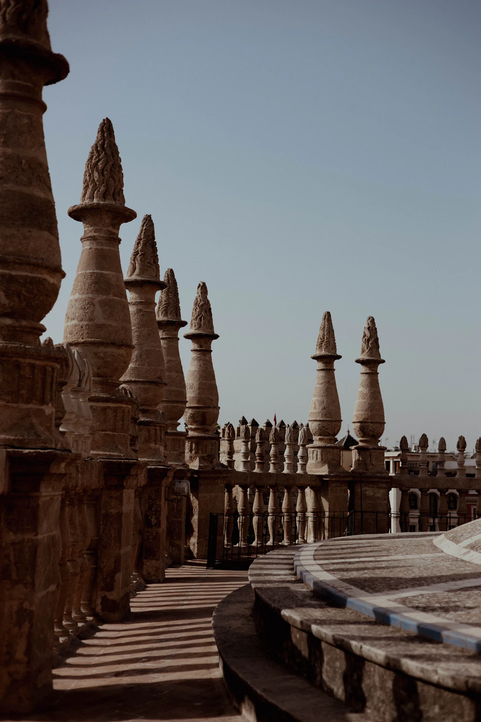 The image depicts a stone structure with multiple spires and columns, situated on a rooftop or balcony. The building features a curved roof that adds to its architectural charm. The perspective is from below, looking up at the imposing height of the structure. The sky above is clear blue, providing a beautiful backdrop for the stone edifice.