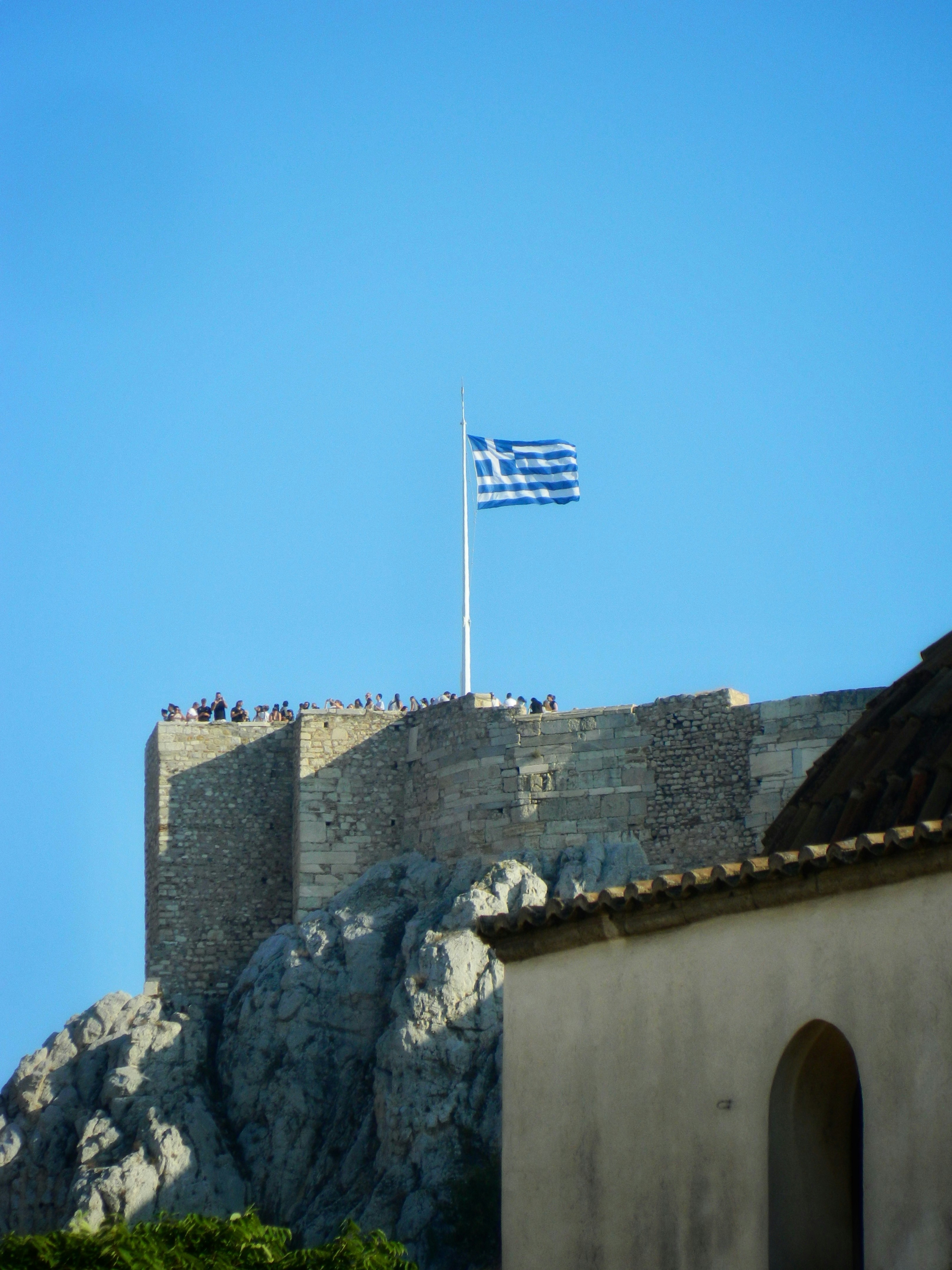 Greek flag flies atop ancient stone fortress under clear sky