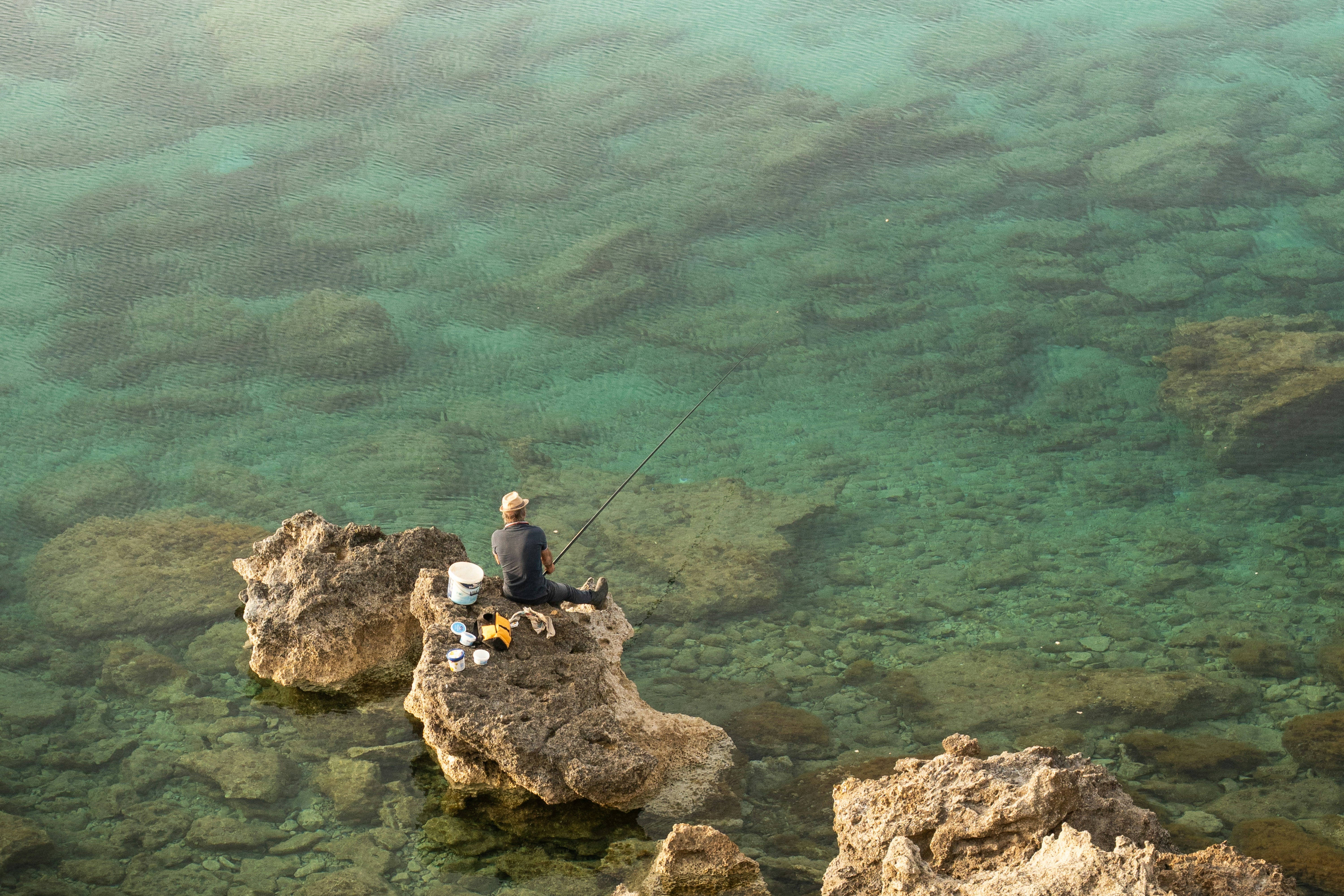 Eine Person sitzt auf Felsen und angelt im klaren türkisfarbenen Wasser.