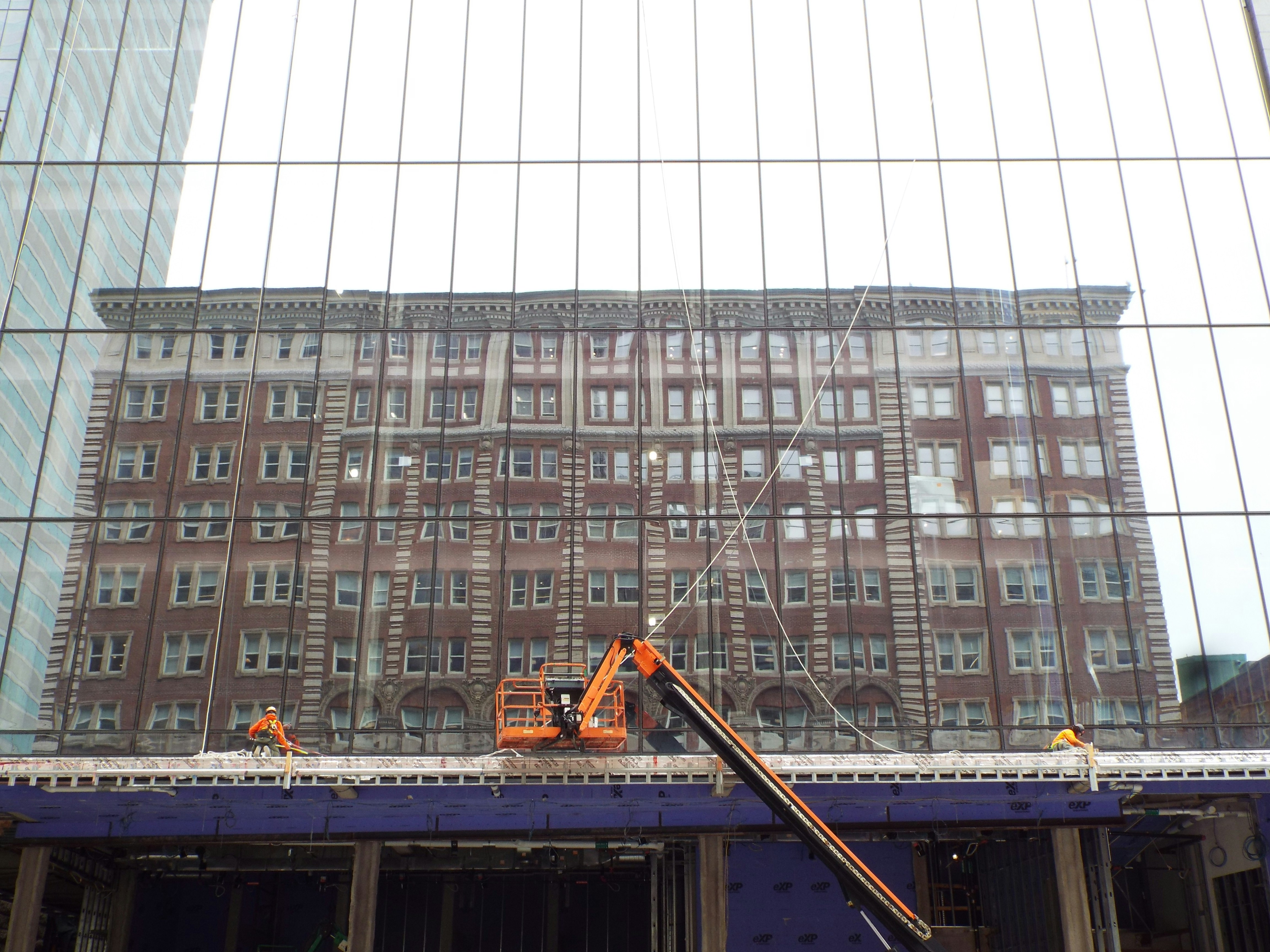 Construction workers on a lift, reflecting the historic architecture of a neighboring building in the glass facade of a modern skyscraper.
