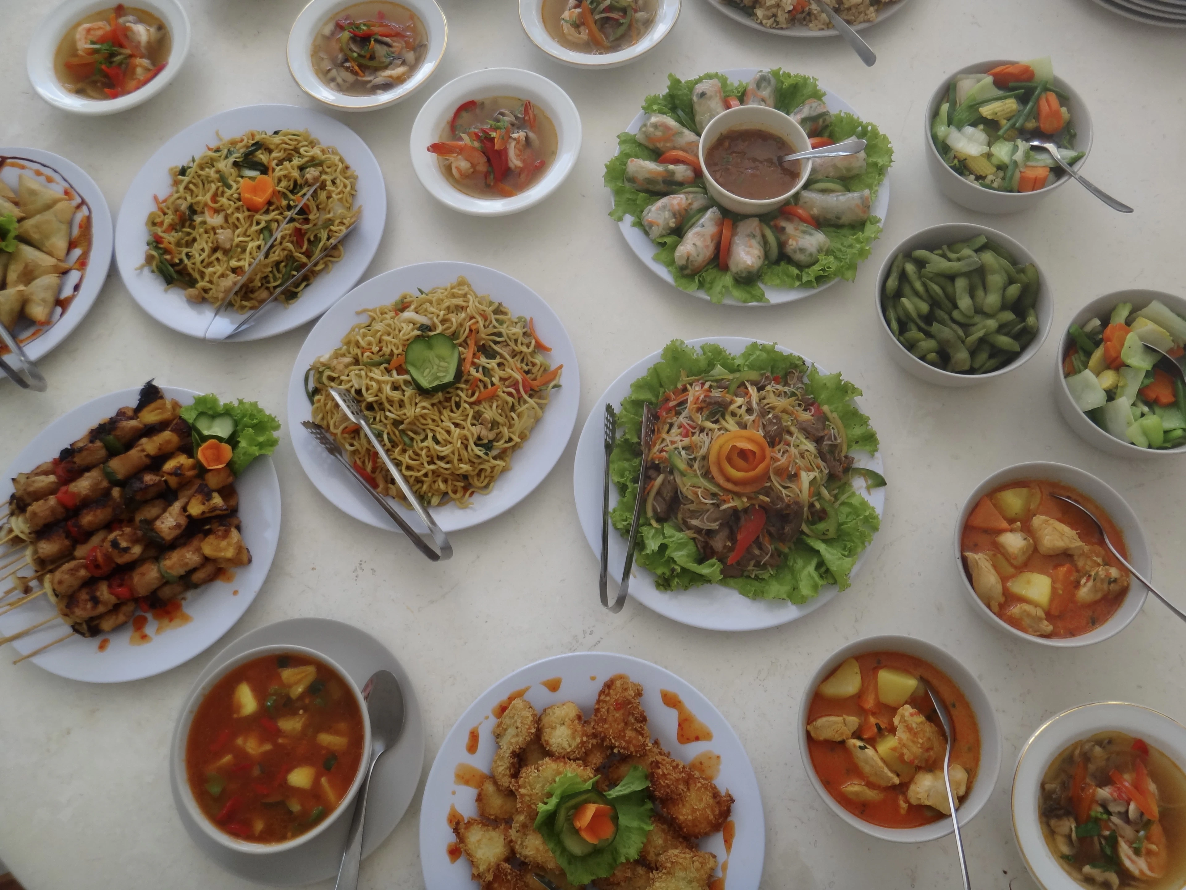 An overhead spread of Southeast Asian dishes on a table: fried rice, noodles, spring rolls, satay skewers and soups.