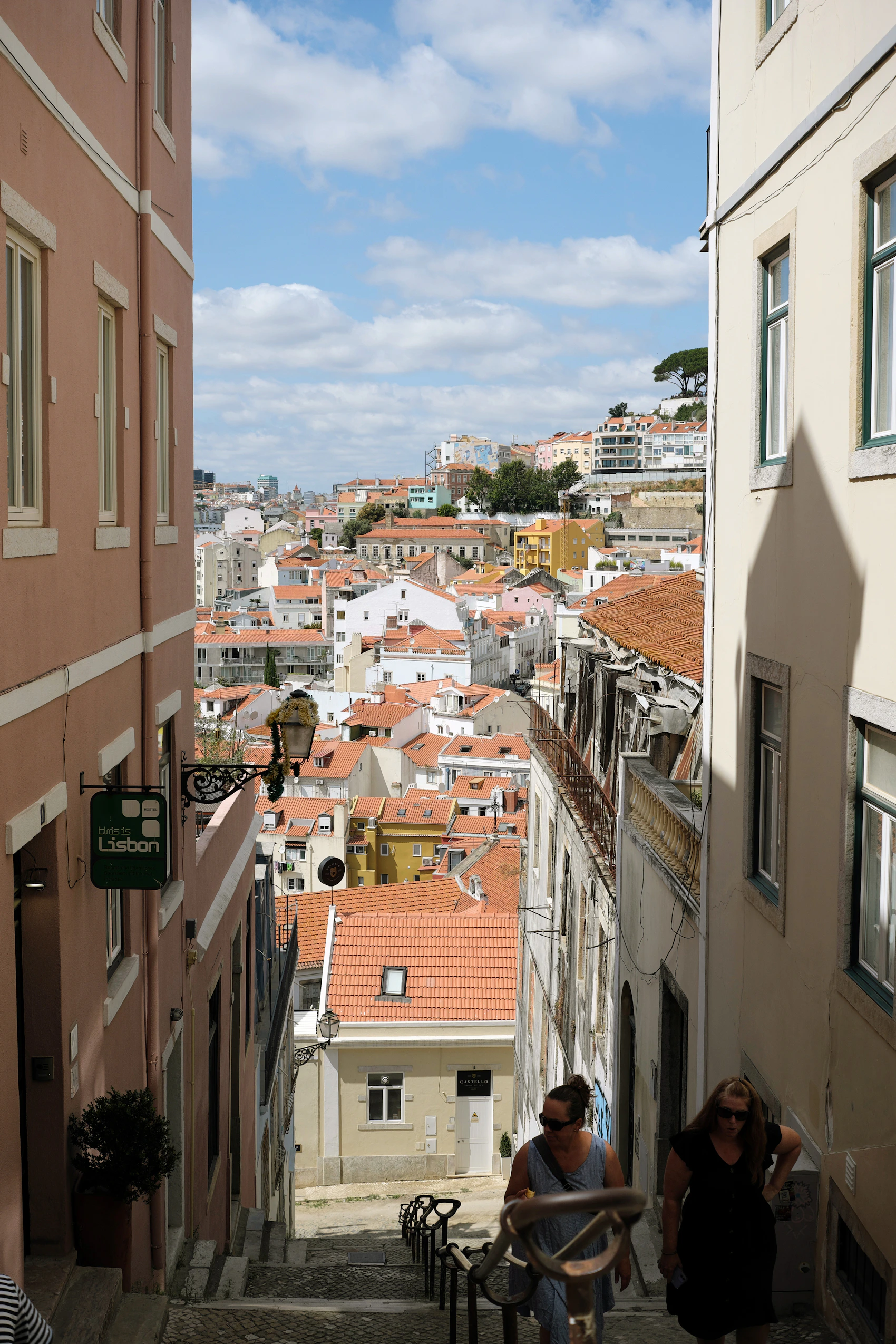 People walking up a steep, narrow cobbled street between Lisbon's tiled roofs