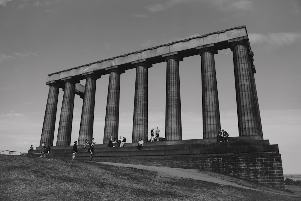 Large stone monument stands on hillside with six tall columns, surrounded by people