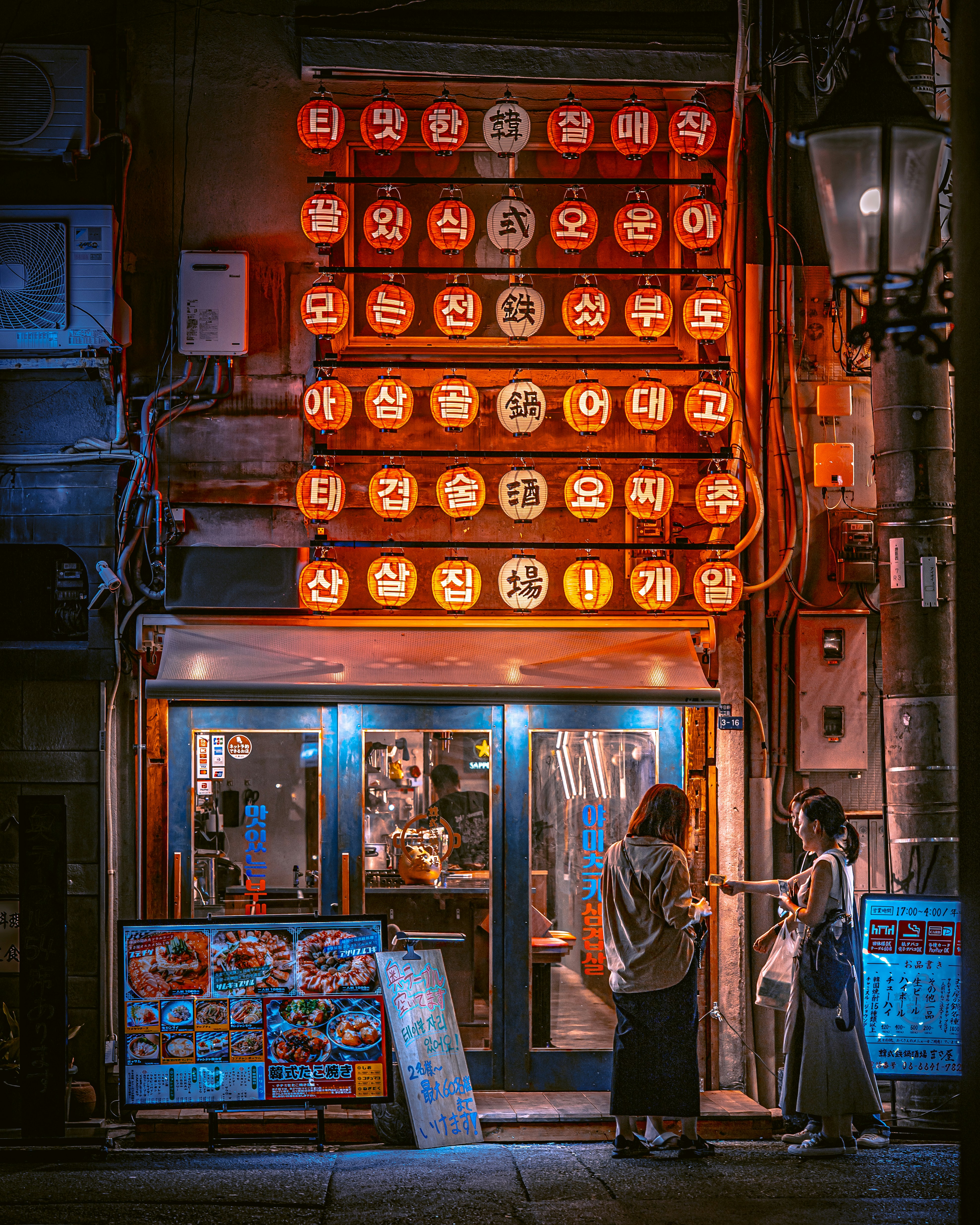 Dos mujeres se paran afuera de un restaurante coreano brillantemente iluminado por la noche.