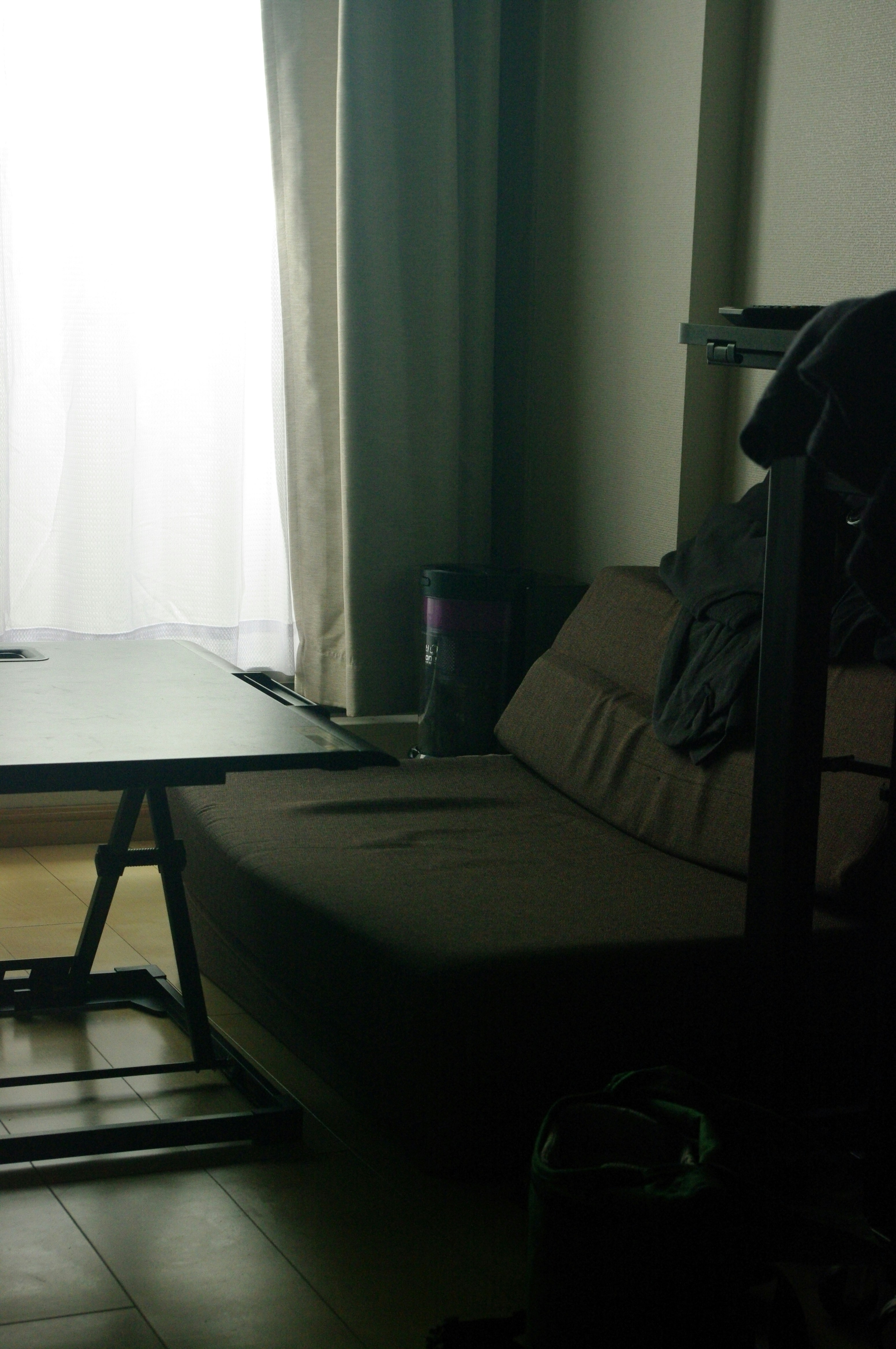 A simple living room corner with a sofa and table is softly lit by daylight through sheer curtains.