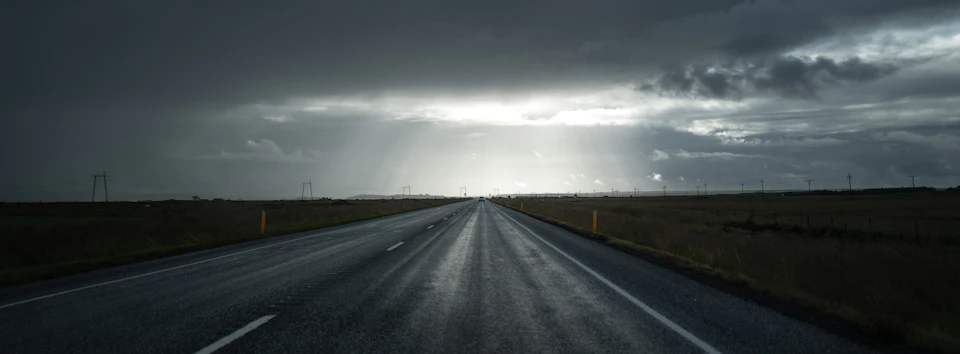 Long, straight road stretches into distance under dark cloudy sky with power lines parallel