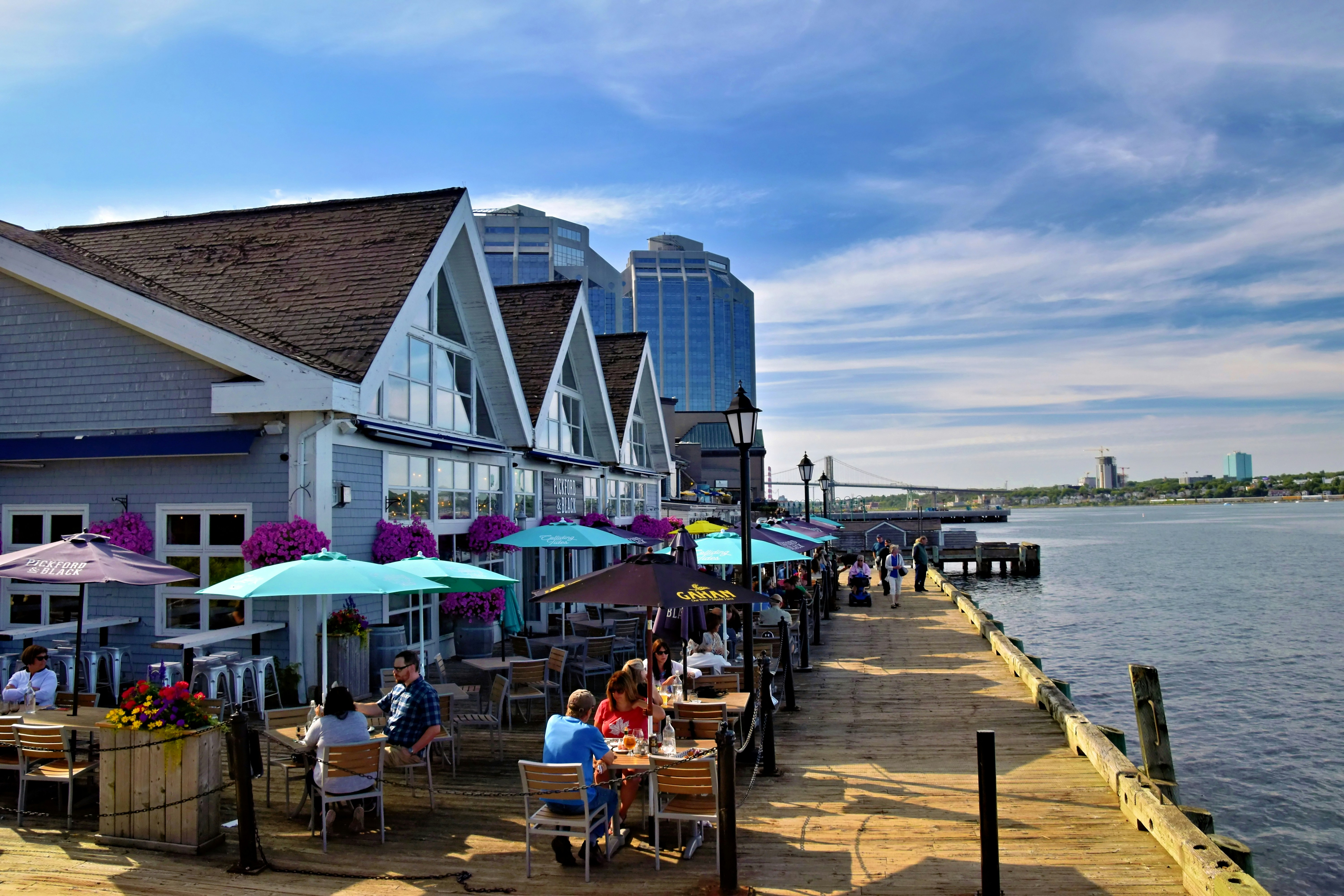 People dining outdoors on a sunny waterfront boardwalk.