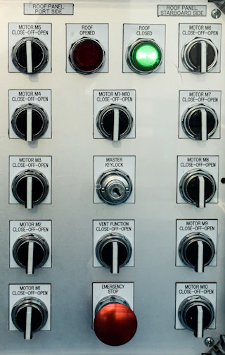 A gray industrial control panel crowded with selector switches, indicator lights, and a red emergency stop button.