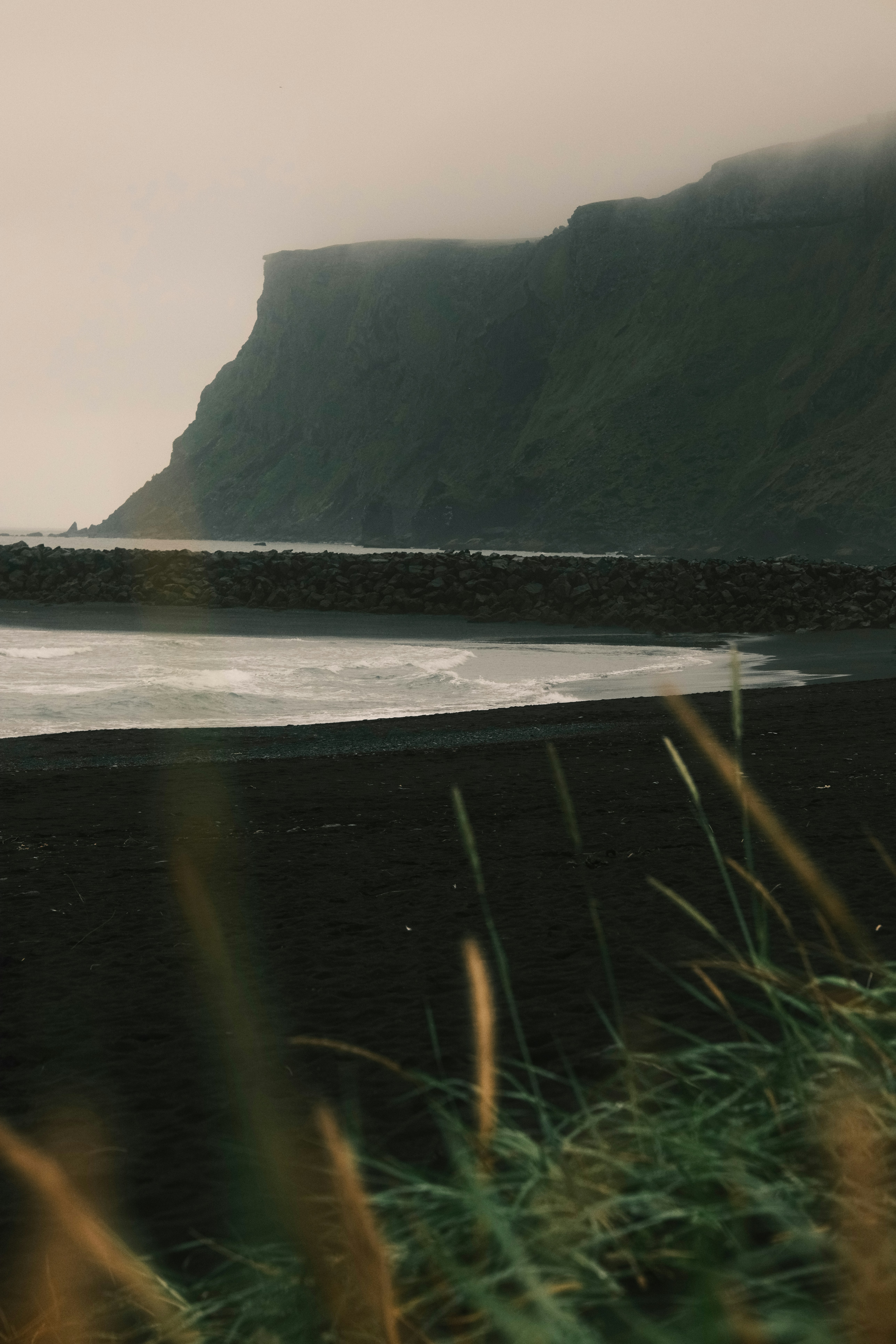 Misty cliffs overlook a dark sandy beach and ocean.