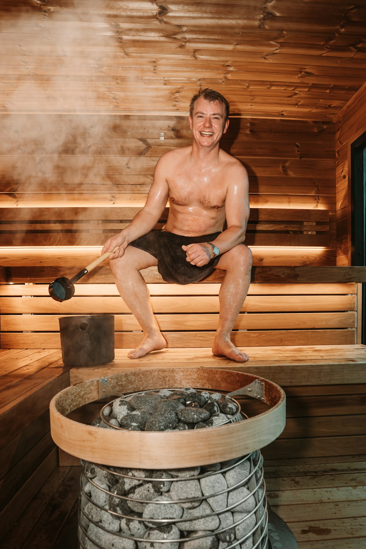 A smiling man in a towel sits on a sauna bench holding a ladle over a bucket, with a round wood-and-stone heater in the foreground and steam rising behind him.