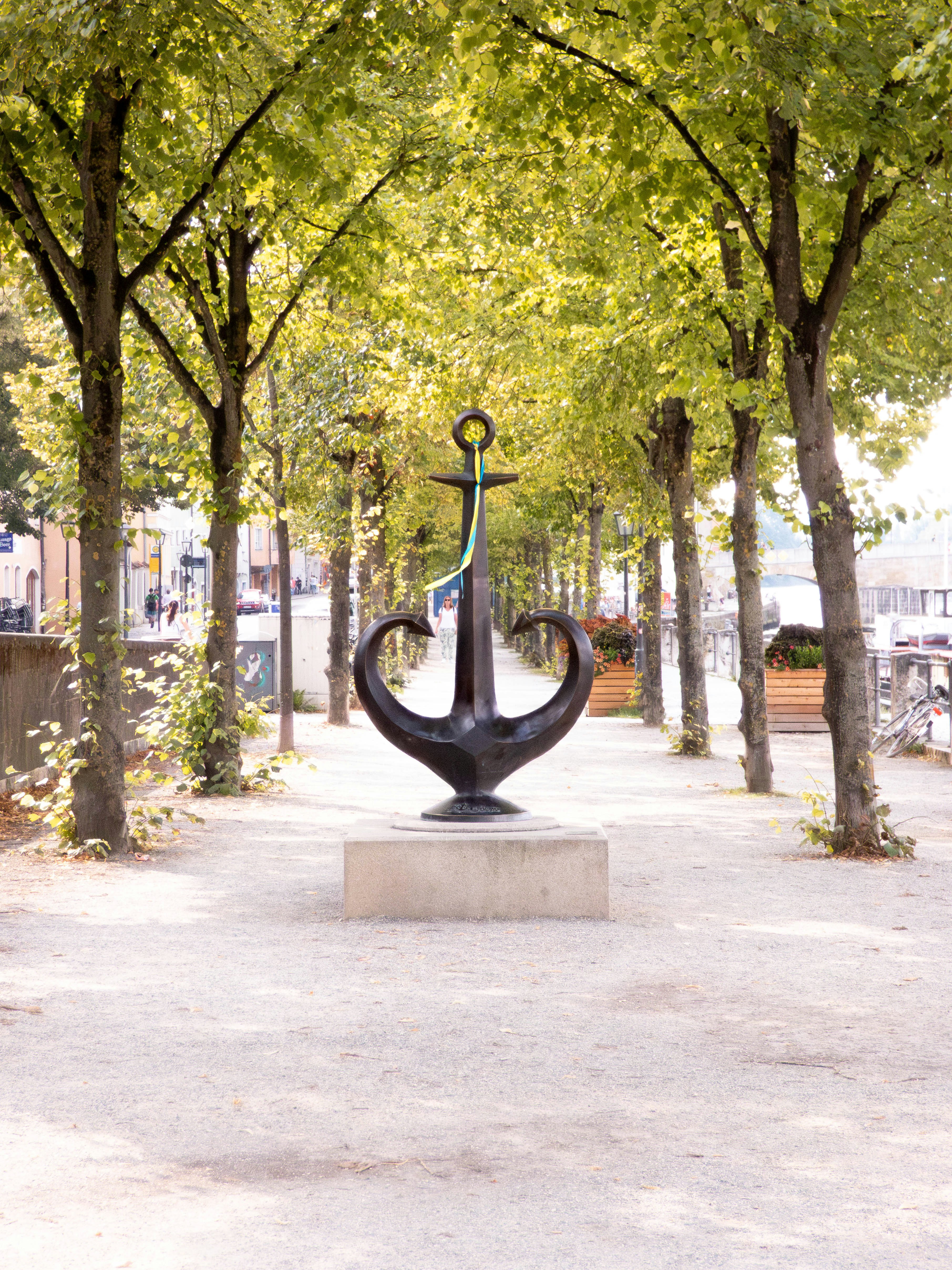 A metal anchor sculpture stands on a pedestal in the middle of a tree-lined pedestrian walkway in Regensburg, Germany.