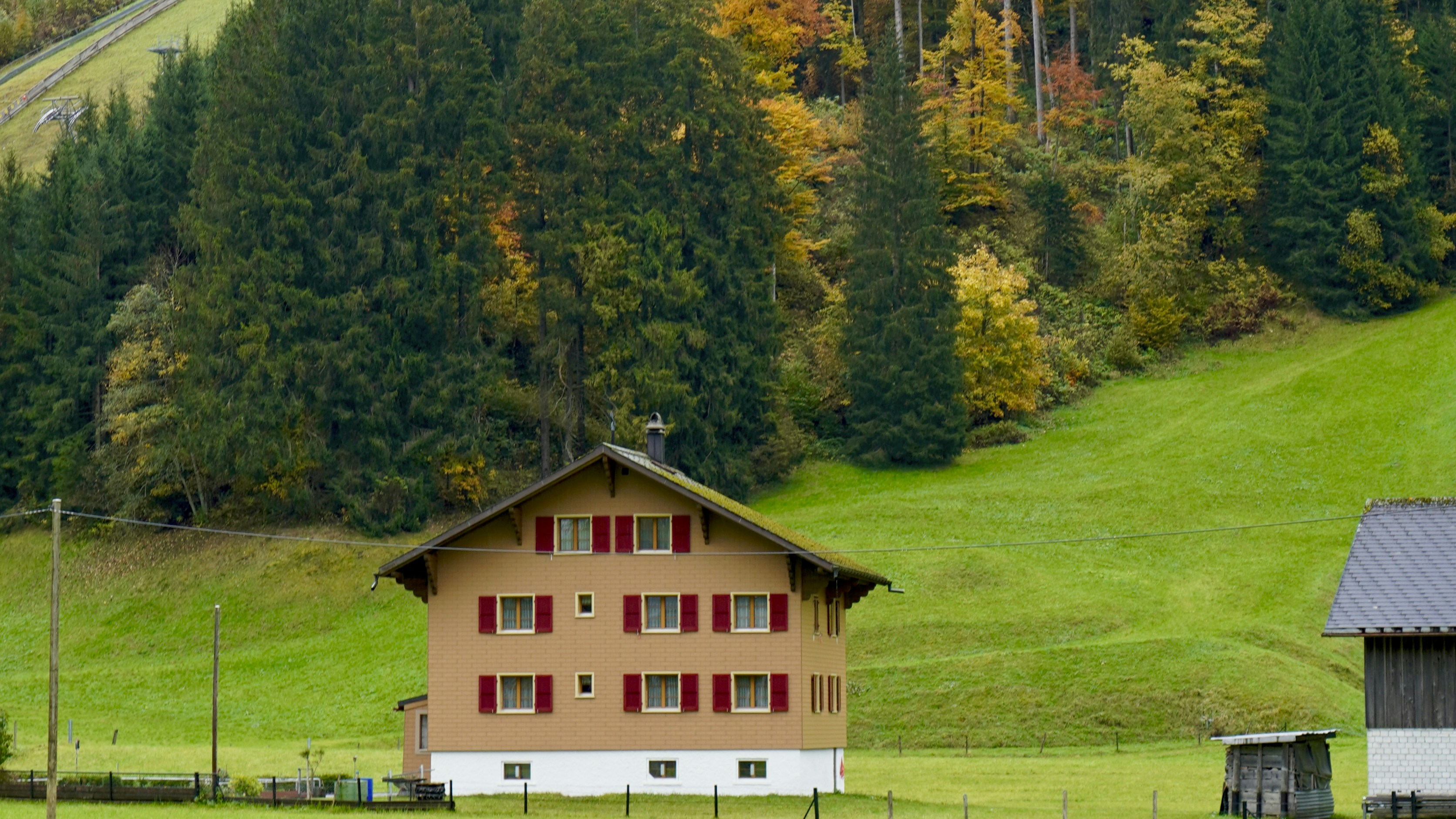 A rustic house sits in a green meadow with autumn trees.