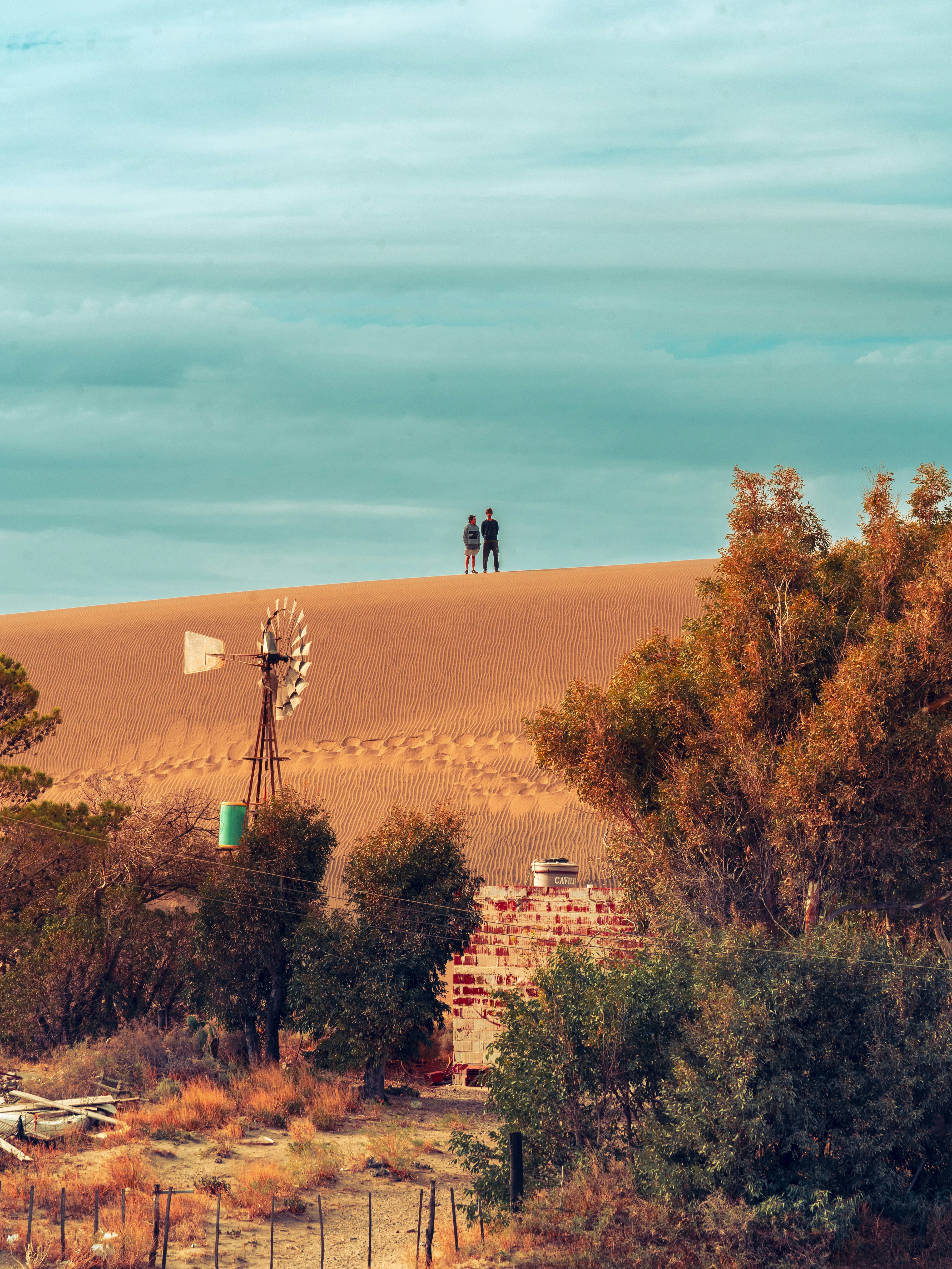 Two people stand atop a sand dune with a windmill.