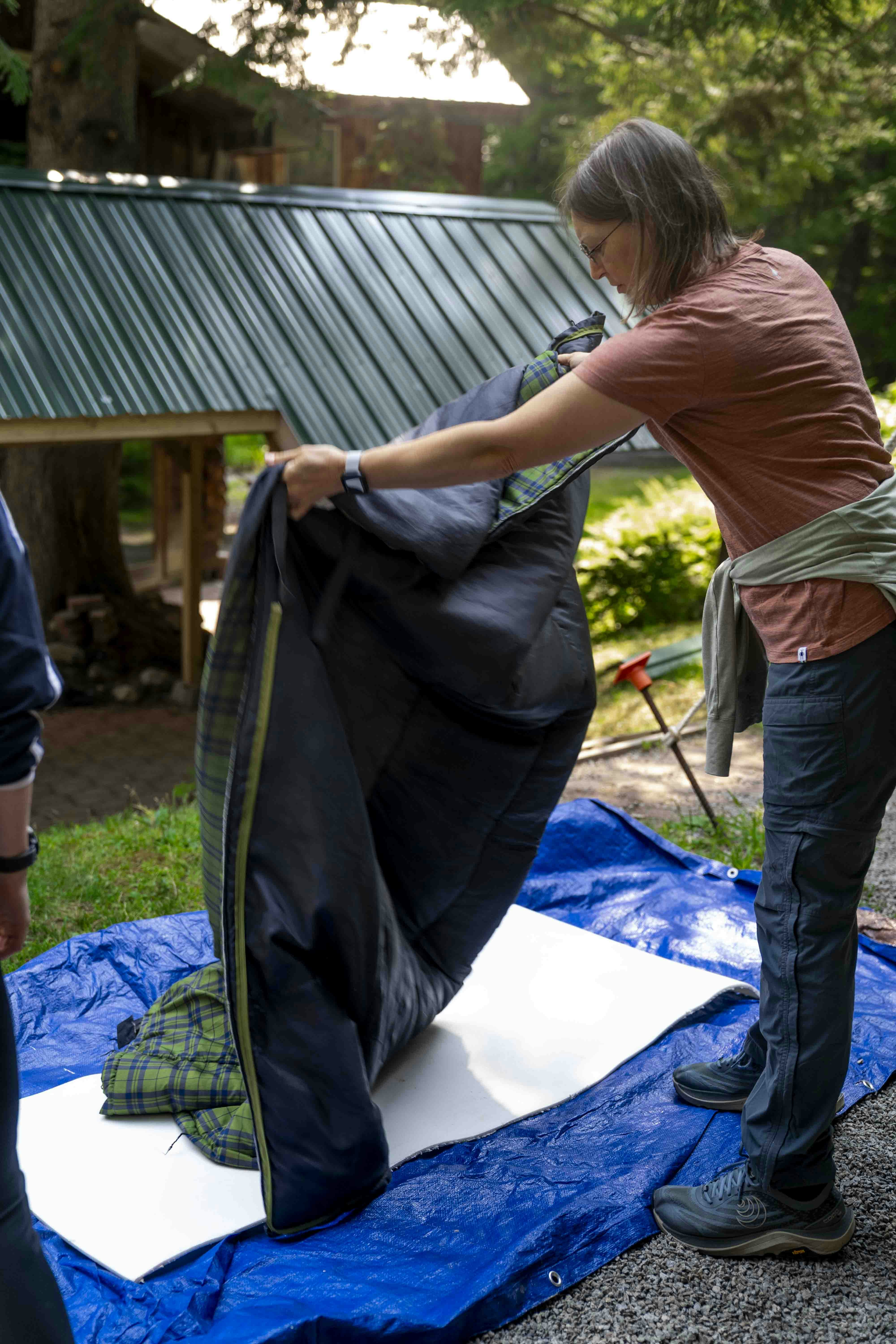 Woman at a campsite shaking out a dark sleeping bag over a blue tarp and foam pad, with trees and a green-roofed shelter behind