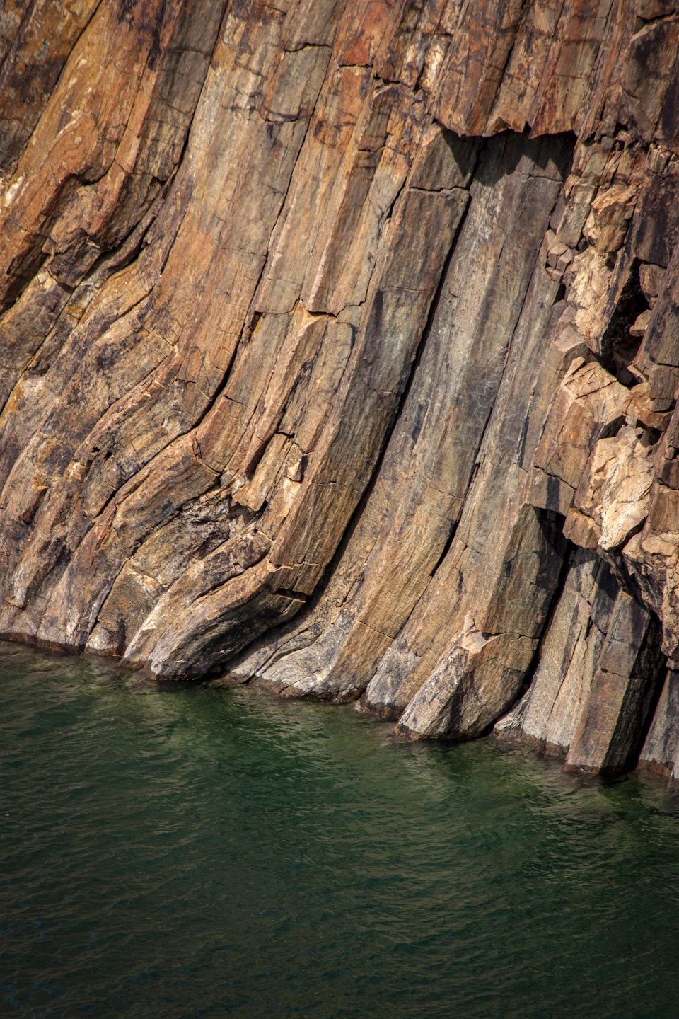 The image depicts a large, jagged cliff face composed of dark brown and gray rock layers. The cliff is situated on the side of a body of water, with the greenish-blue waters reflecting the surrounding landscape. The perspective of the photo is taken from below, looking up at the imposing height of the cliff.