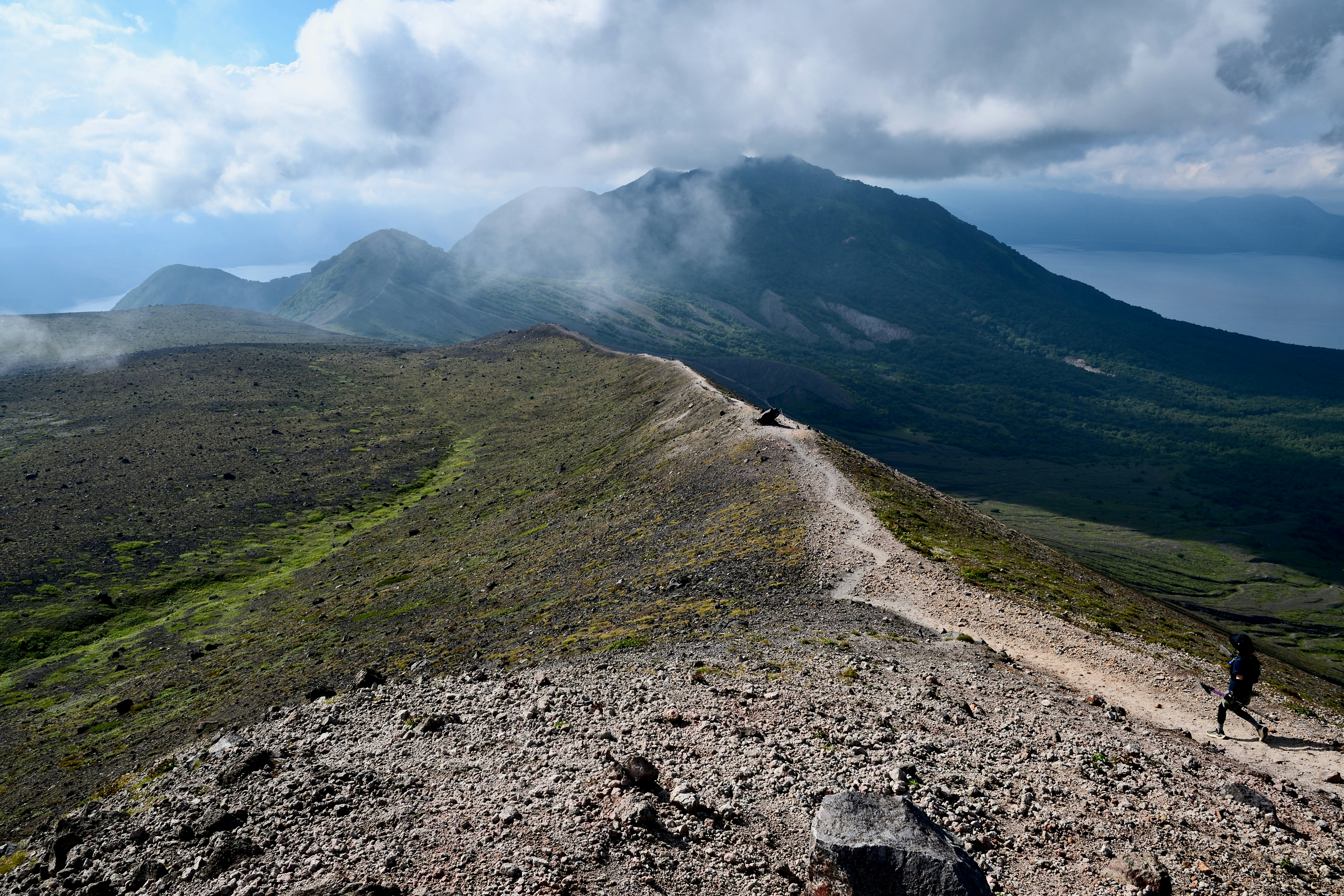 A person hikes a rocky mountain trail with clouds.