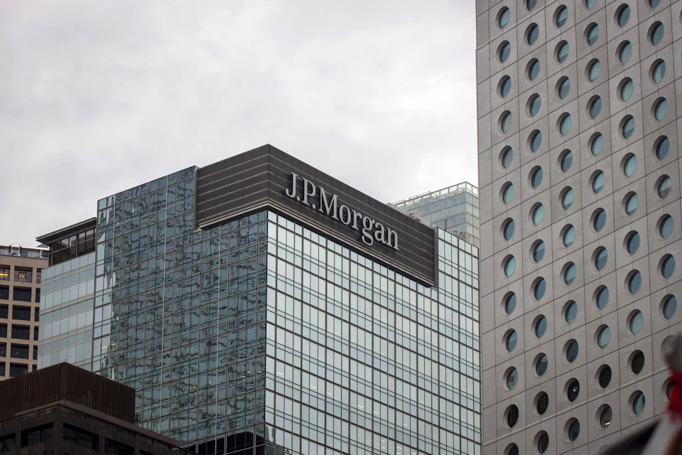 The image shows a tall, modern glass and steel building with a large J.P. Morgan sign on its side. The building is situated between two other structures, one gray and one white, creating an urban skyline. The sky above appears overcast, casting a soft light over the scene.