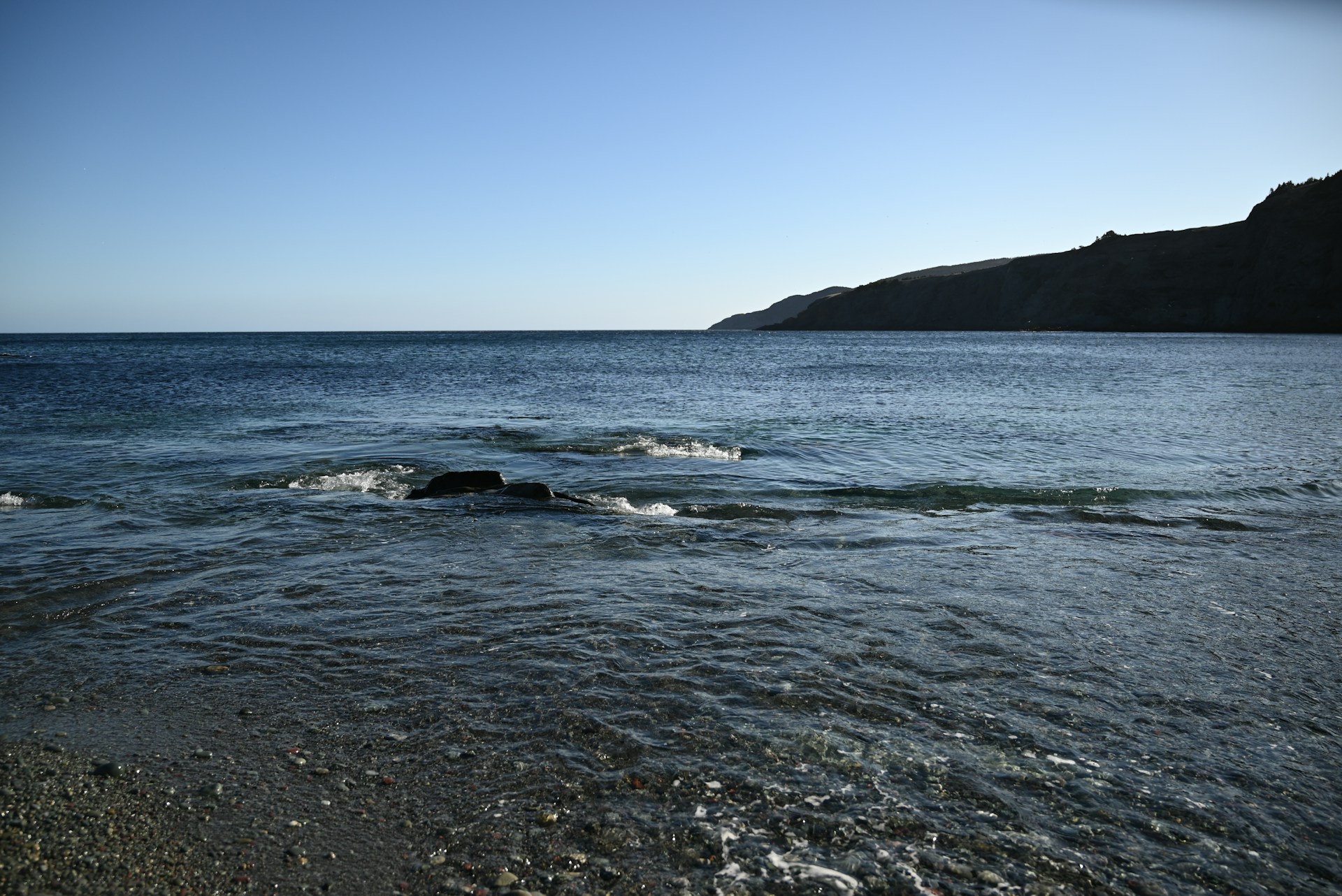 Calm ocean waves gently lap a pebble beach under clear sky.