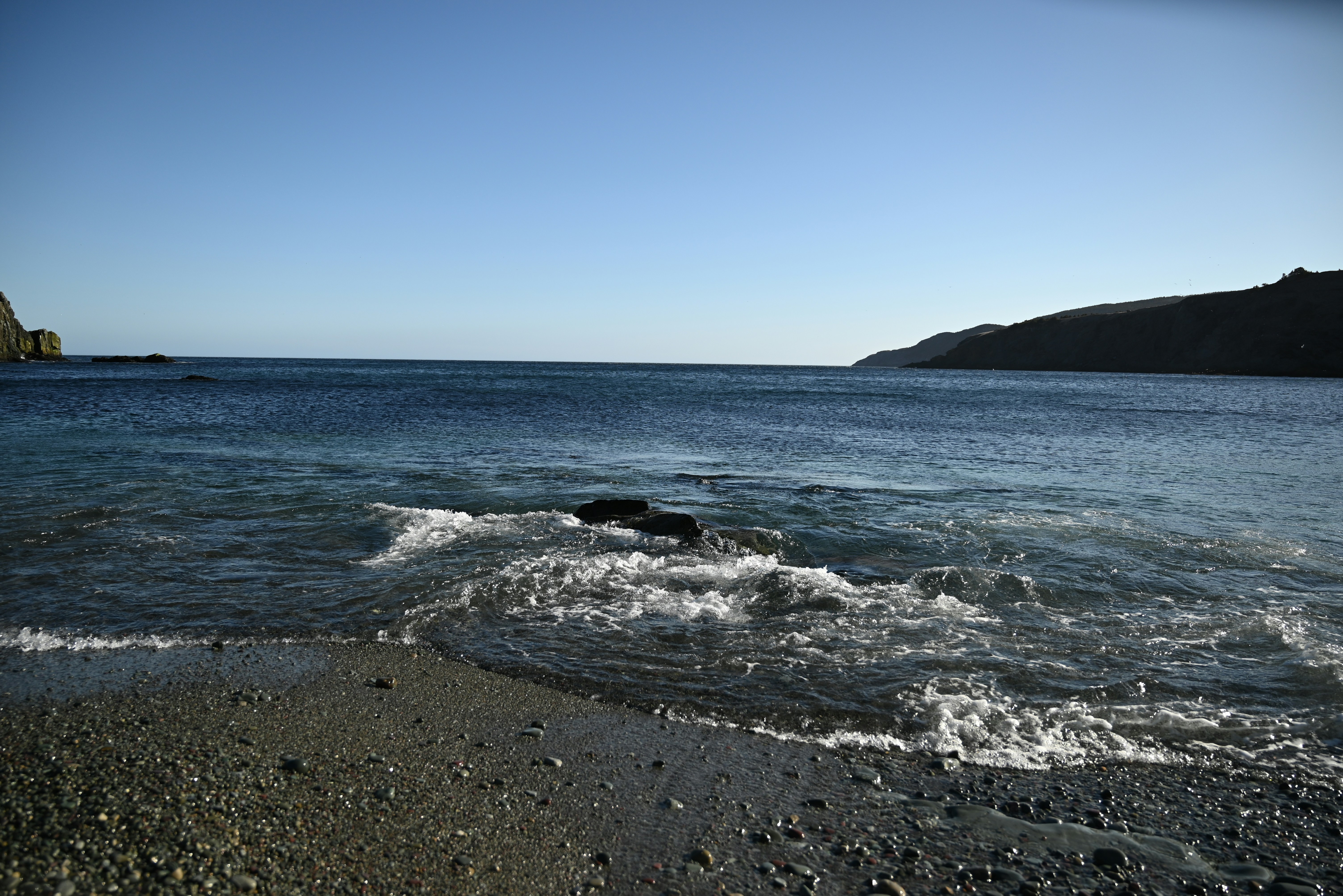 Les vagues s’échouent doucement sur une plage de galets sous un ciel dégagé.