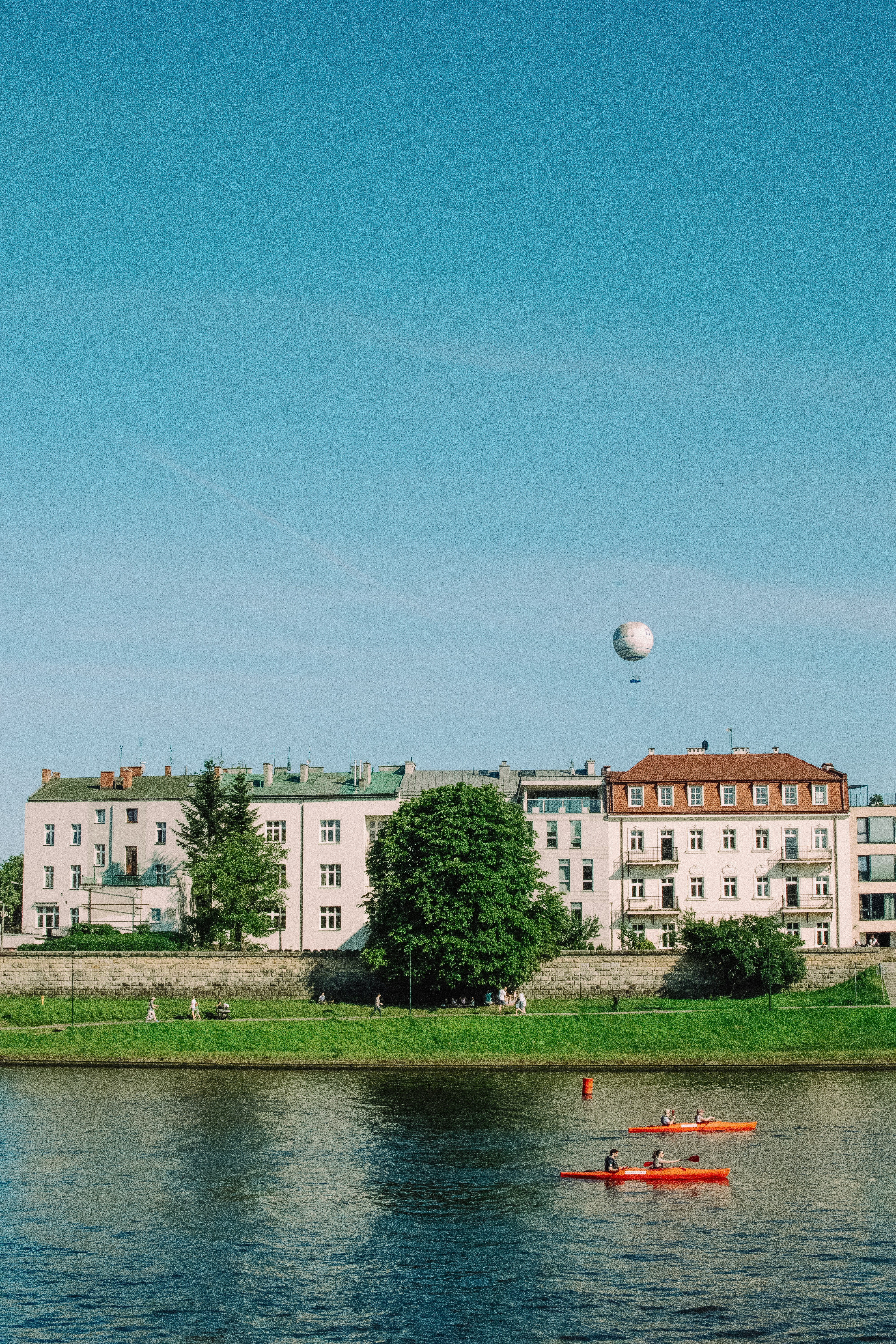 Kayakers on a river with buildings and hot air balloon.