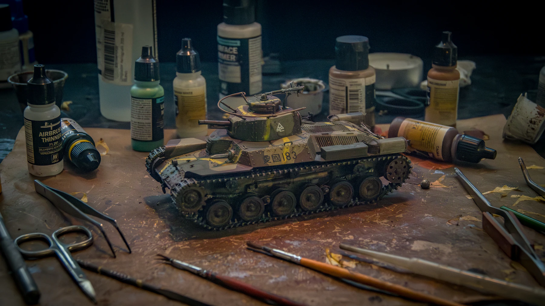 A finished camouflaged tank model on a workbench, surrounded by paint bottles, brushes and tools