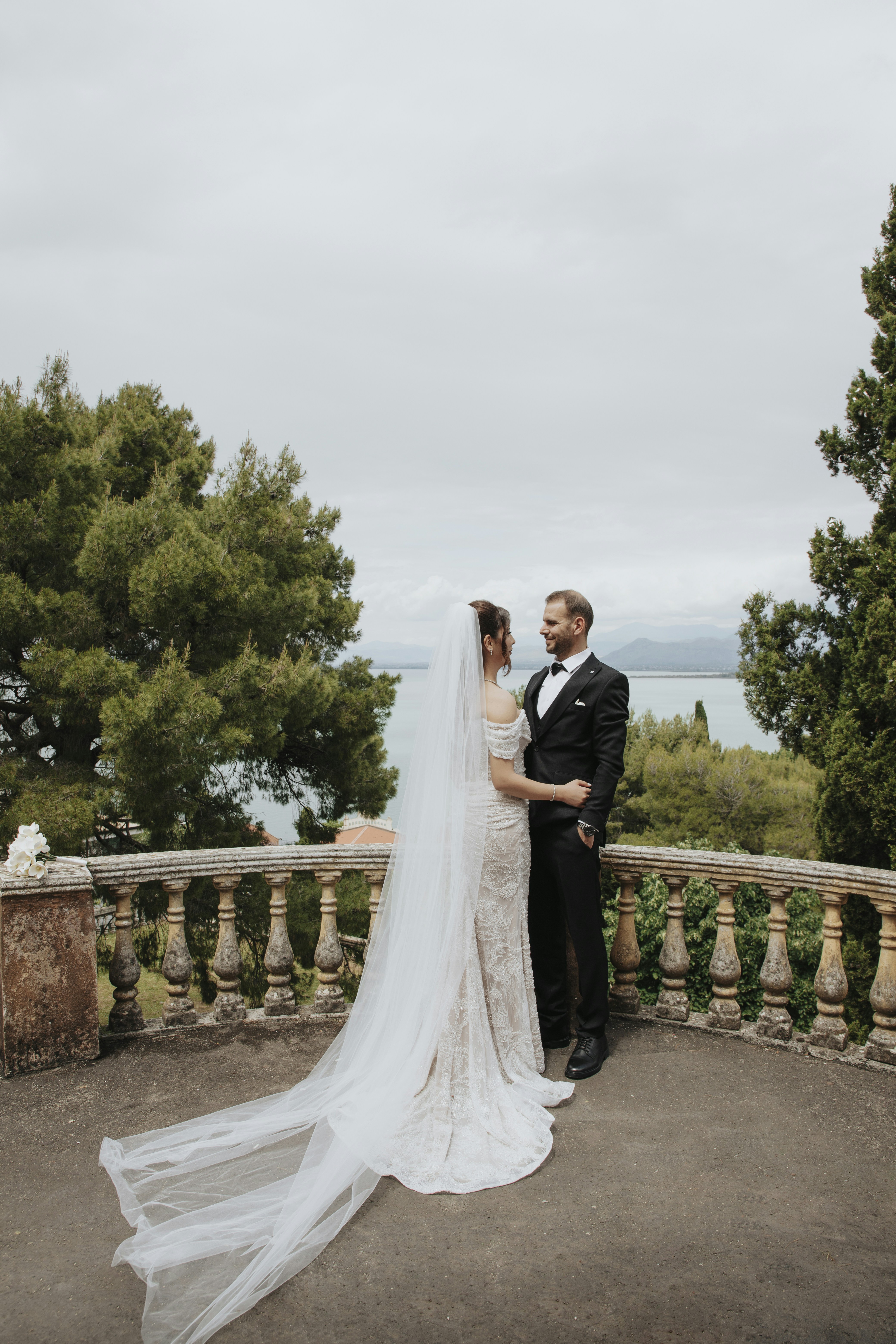 A bride and groom stand on a balcony overlooking water.