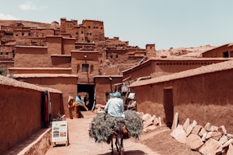 Man carries large bundle of hay on bicycle through village.