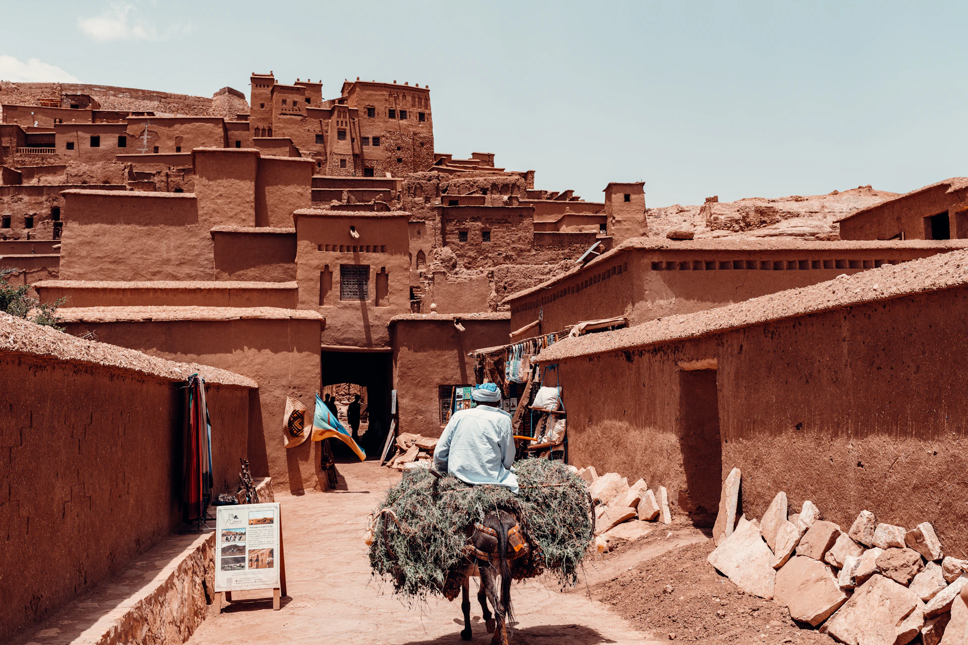 Man carries large bundle of hay on bicycle through village.