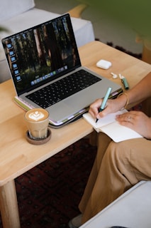 Person writing in notebook next to laptop and coffee.