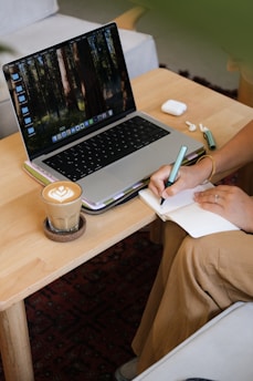 Person writing in notebook next to laptop and coffee.