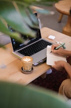 Person writing in notebook next to laptop and coffee.