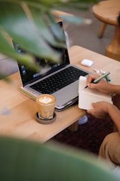 Person writing in notebook next to laptop and coffee.