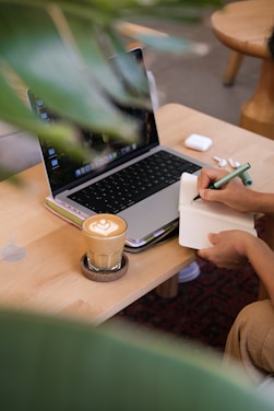 Person writing in notebook next to laptop and coffee.