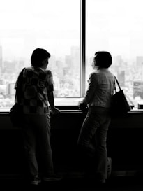 Two people look out a large window at city.