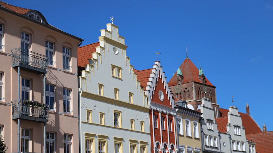 The image depicts a row of old buildings with red roofs, standing tall against a clear blue sky. The buildings are painted white and yellow, creating a striking contrast that highlights their architectural details. Each building features a balcony on its second floor, adding to the charm of the scene. The perspective of the photo is from ground level, looking up at the buildings, emphasizing their height and grandeur.