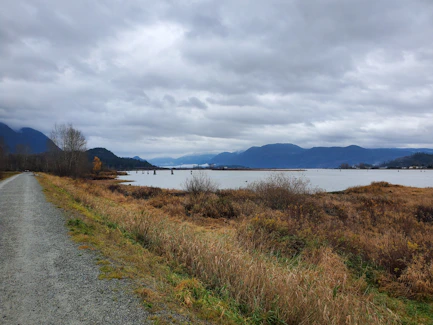Gravel path beside a calm lake with mountains under cloudy sky