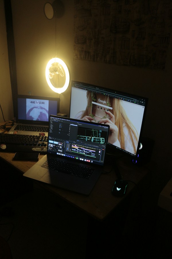 A dim desk lit by a circular ring light where a laptop runs video-editing timeline software while an external monitor beside it displays a young