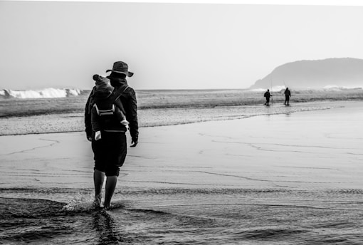 Person with baby carrier walks on a wet sandy beach.