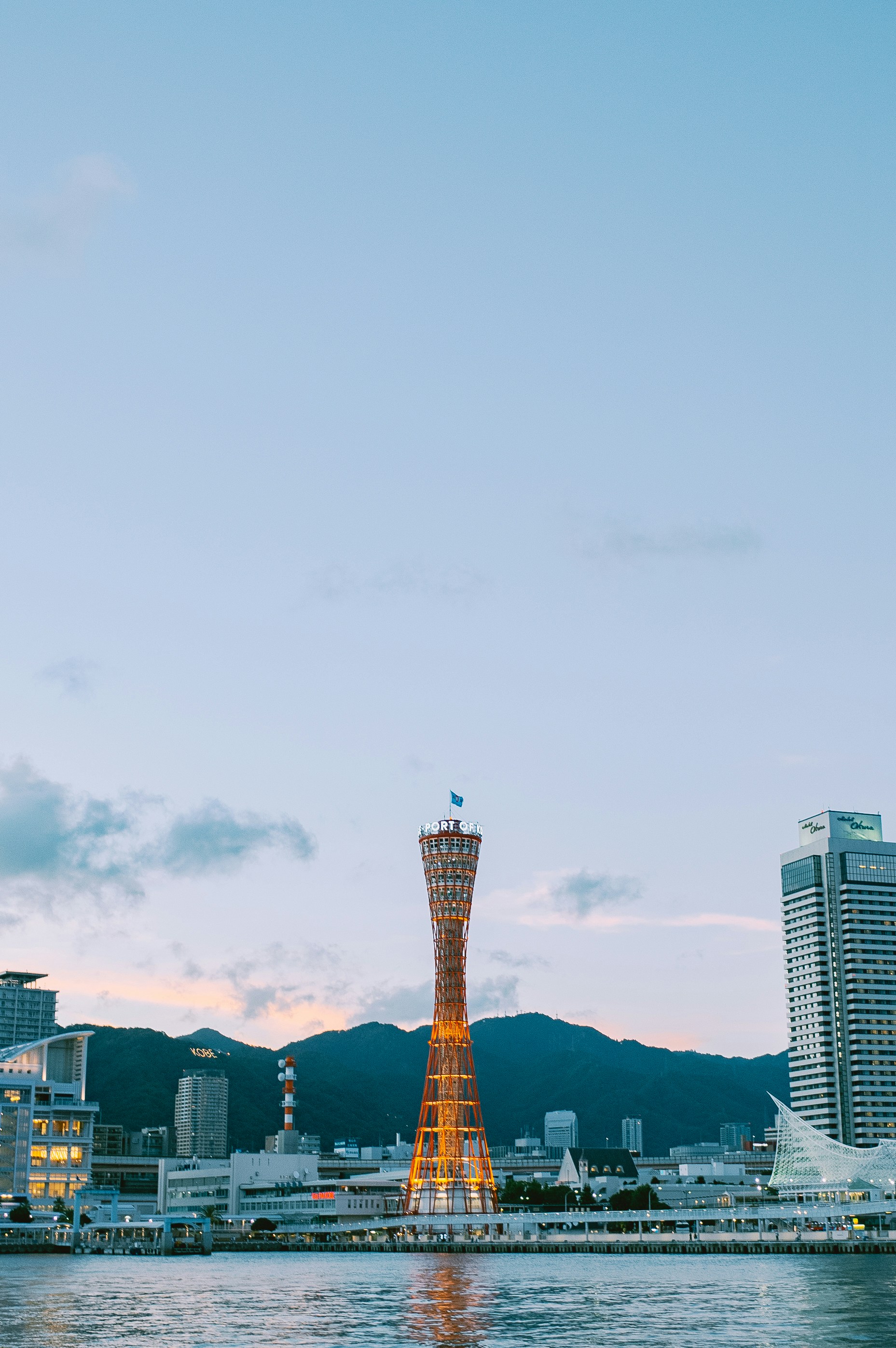 Kobe port tower illuminated at dusk with cityscape and mountains.
