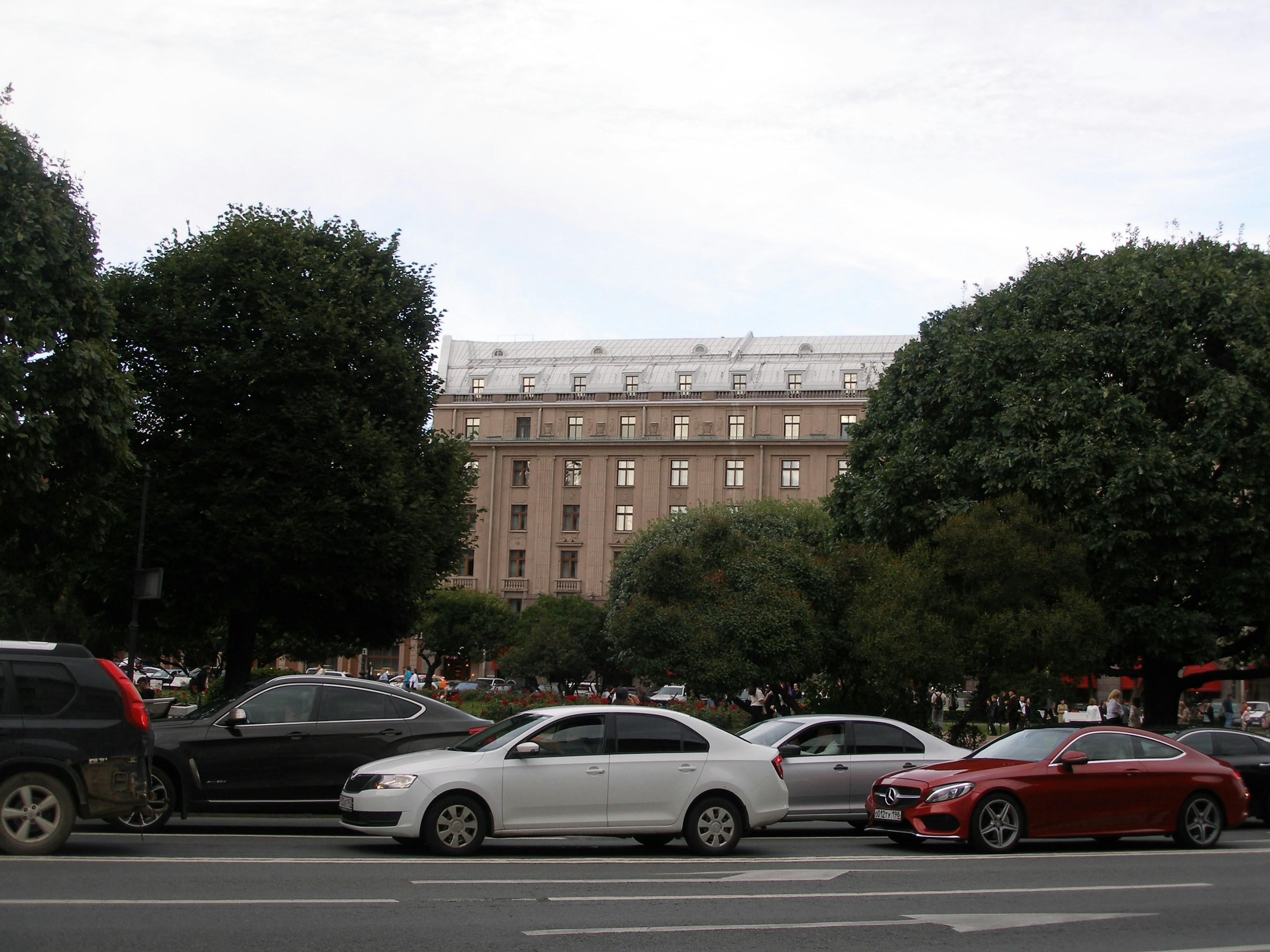 Cars parked on a street with a large building behind trees.