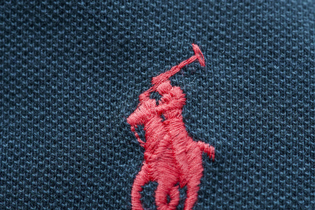 Logo bordir kecil di dada polo pique gelap, tekstur tusukan satin terlihat dari dekat