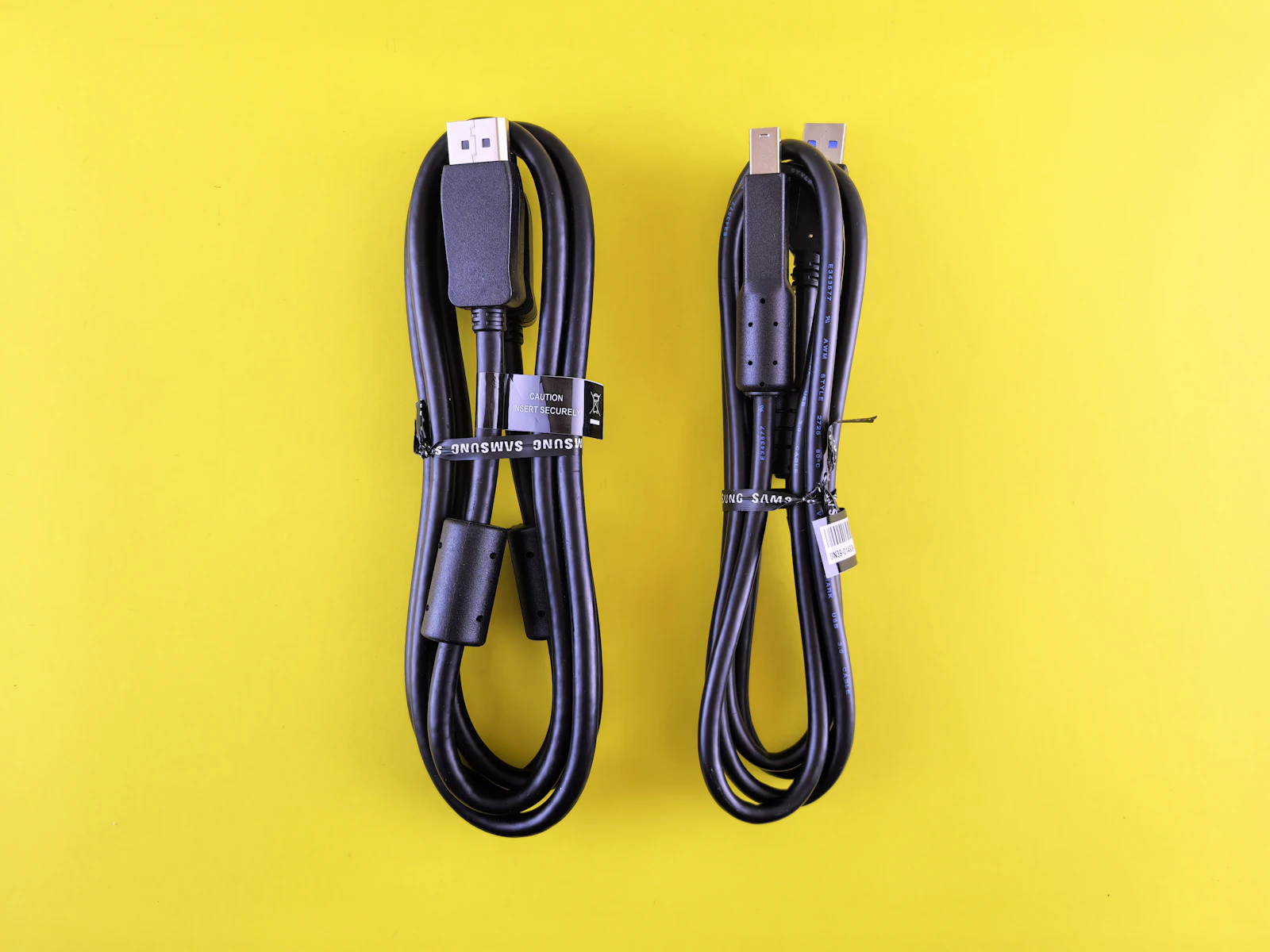 Two coiled black cables on a bright yellow background: a DisplayPort cable with a ferrite bead and Samsung zip tie beside a USB 3 cable with Type-A and Type-B connectors